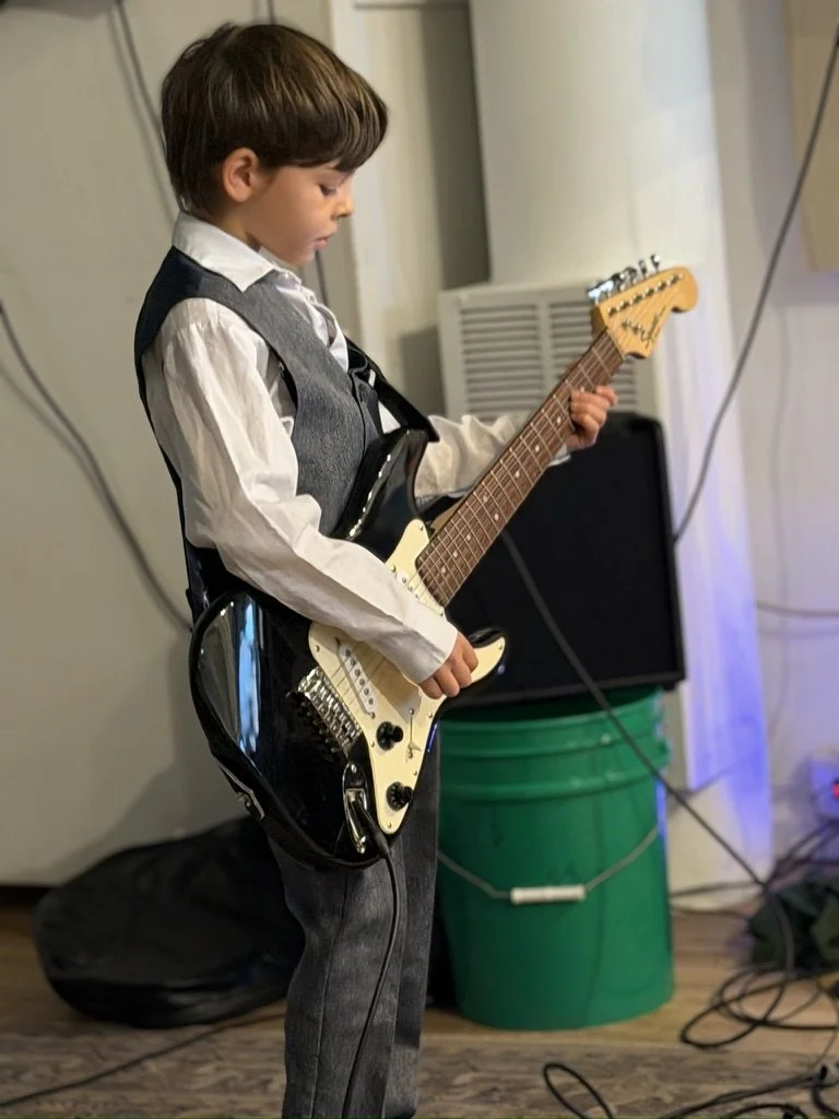 A young boy dressed in a white shirt and gray vest plays an electric guitar.