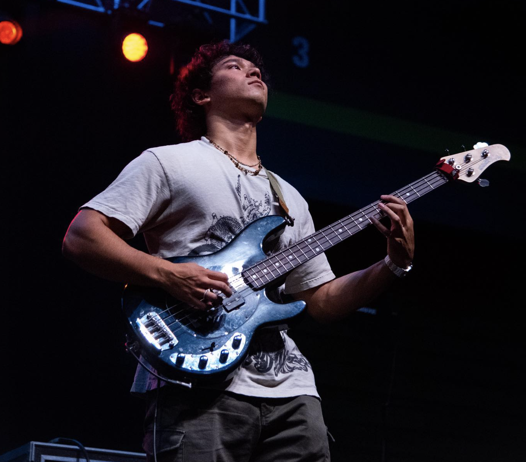 A man with curly hair and a beard playing an electric guitar on stage, illuminated by colorful stage lights with a striped backdrop.