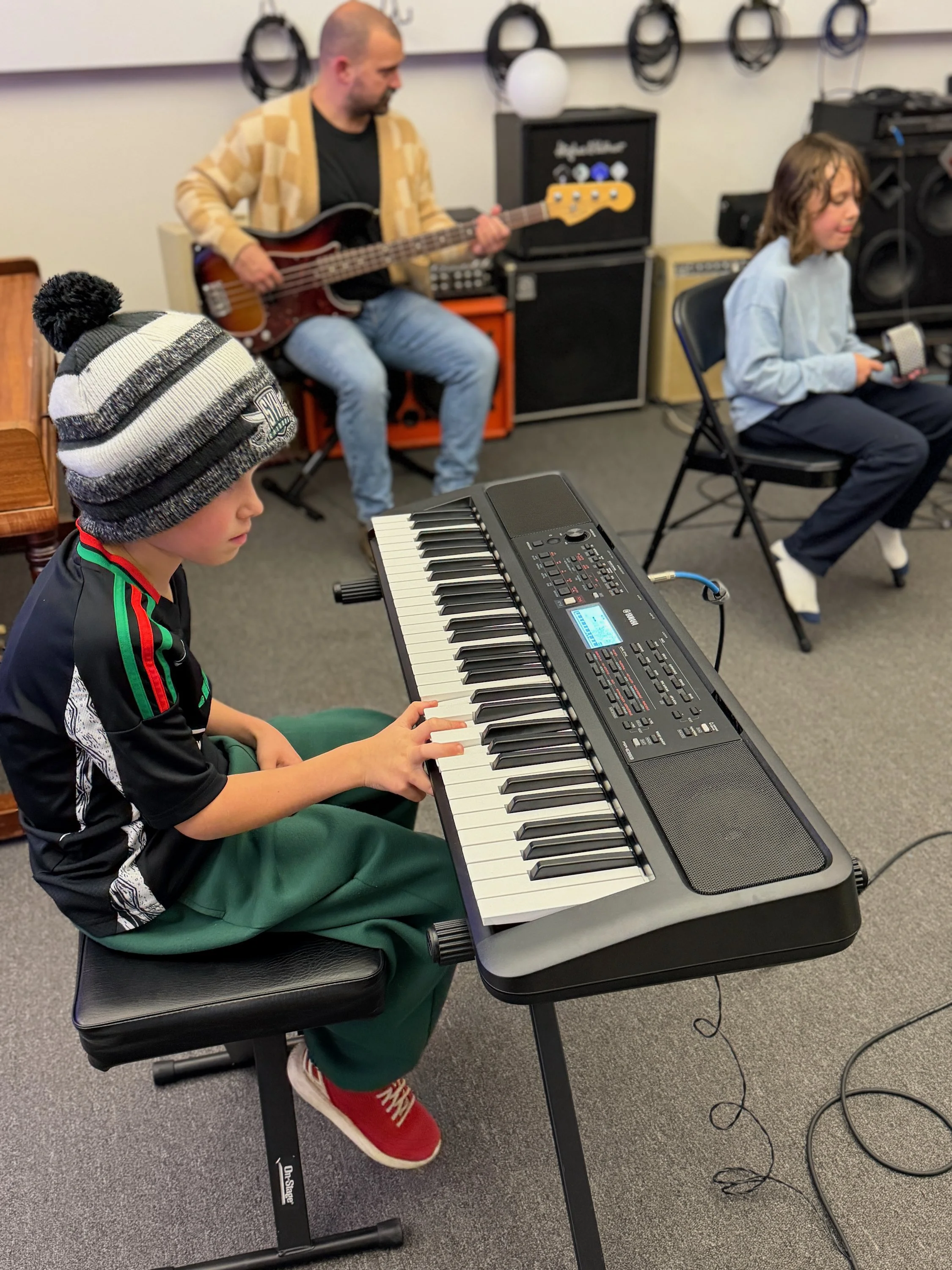A young boy wearing a striped beanie and a sports jersey playing a digital keyboard in a music practice room. In the background, a man with a bald head playing an electric bass guitar, and a girl with long hair sitting on a chair holding a tambourine, all surrounded by musical equipment and amplifiers.