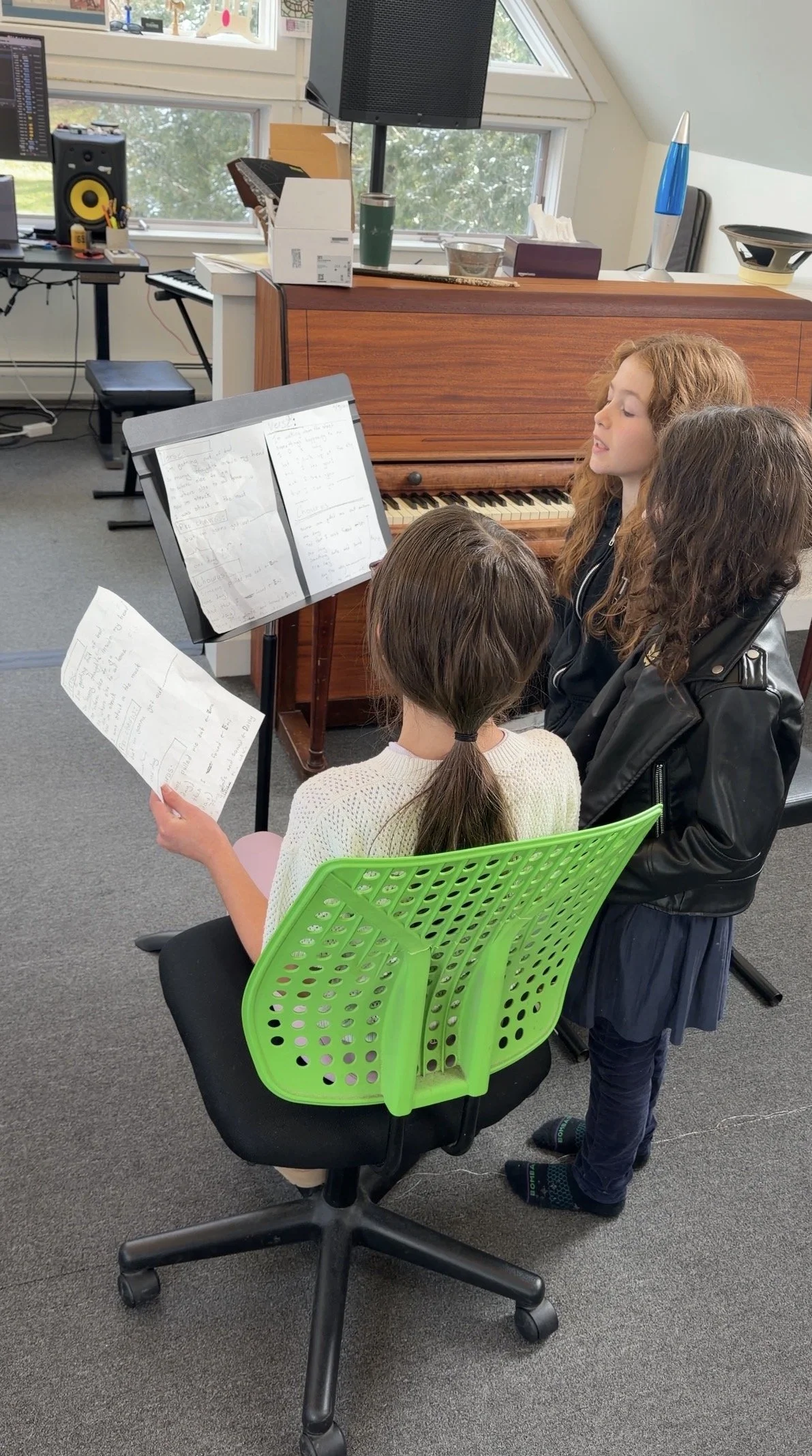 Three young girls are practicing singing in a music room. One girl with long curly red hair is singing and standing next to a seated girl with dark hair in a ponytail. The third girl with long, curly brown hair is standing, holding music sheets, and wearing a black leather jacket. There is a wooden piano behind them, a music stand with sheet music, and a computer and speaker setup in the background.