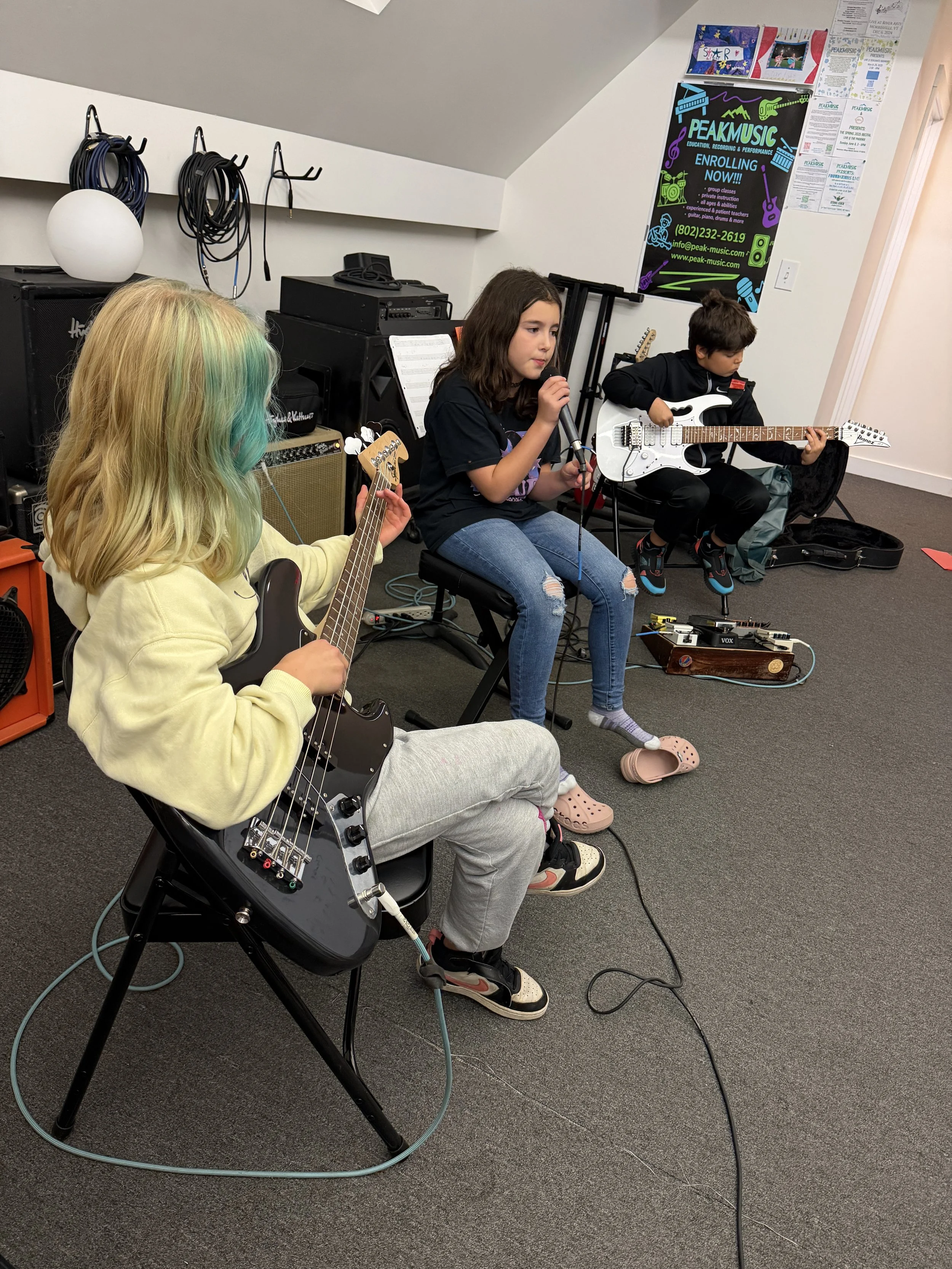 Three children playing electric guitars and singing in a music rehearsal room with equipment, posters, and cables.