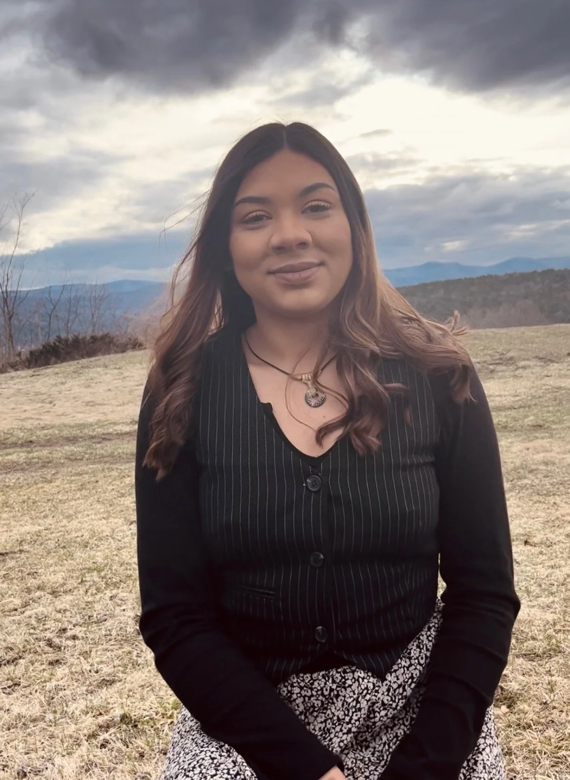 Janéa Hudson with medium-length wavy hair smiling outdoors in a field with mountains and cloudy sky in the background.