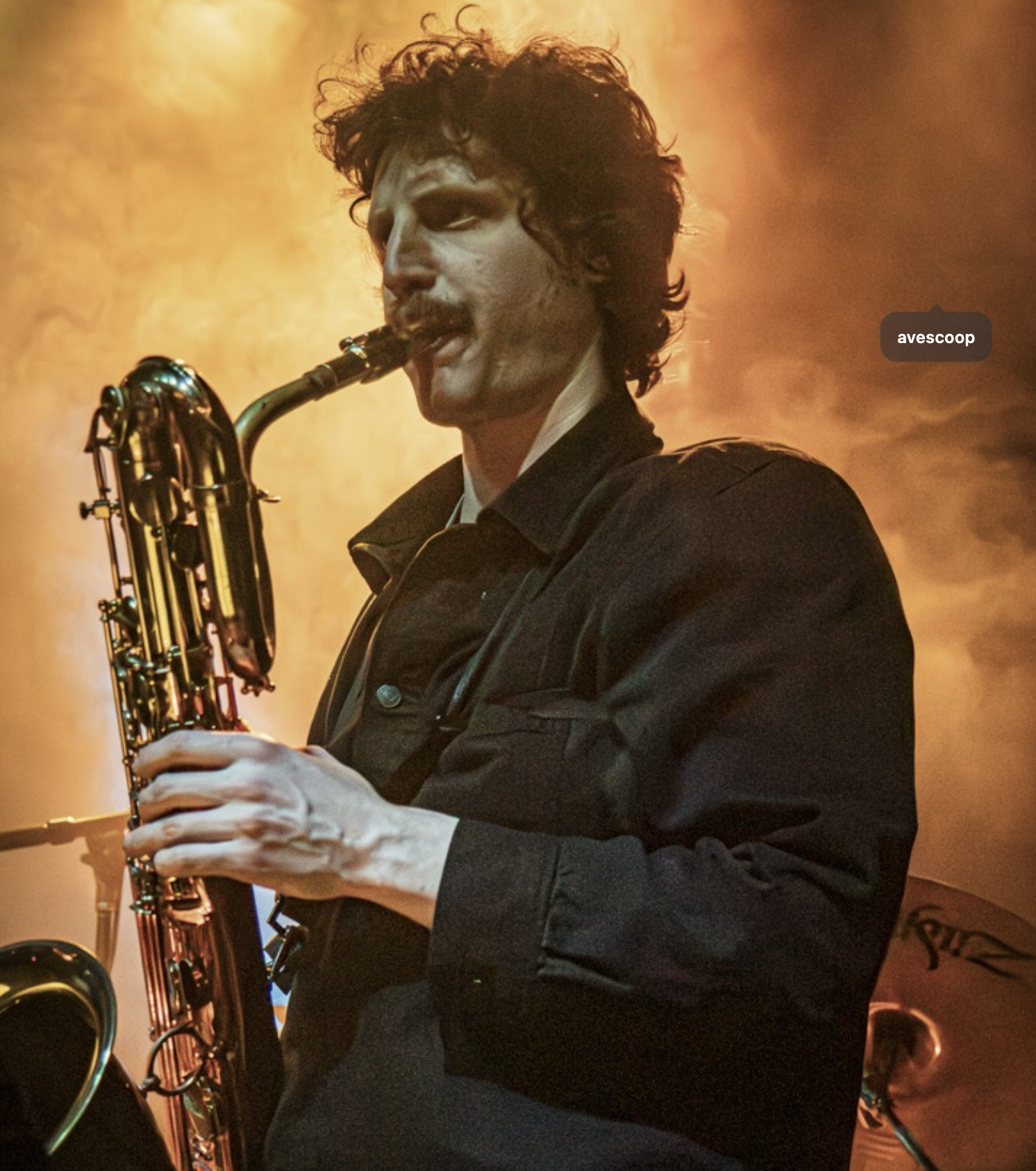 A man with long brown hair wearing a striped navy blue and gray shirt, holding a clarinet, smiling in a music studio surrounded by musical equipment and framed pictures.
