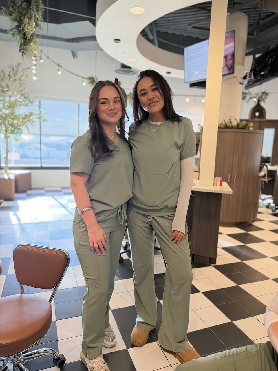 Two women wearing matching green medical scrubs stand side by side inside a bright salon or clinic space with tiled floors, styling stations, and large windows letting in natural light.