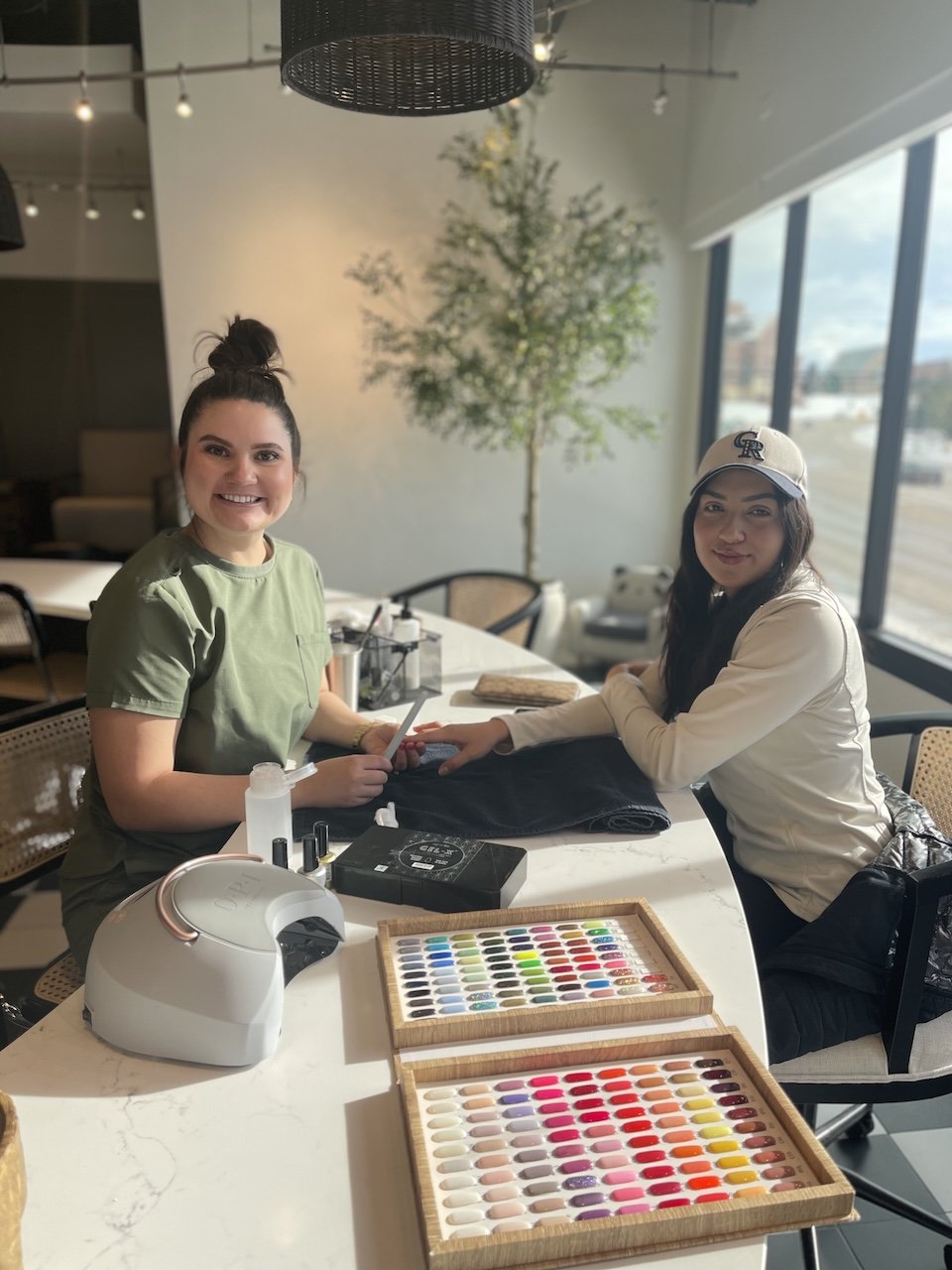 Two women sit at a nail salon table; one is a nail technician working on the other's nails. Nail polish color samples are displayed on the table.