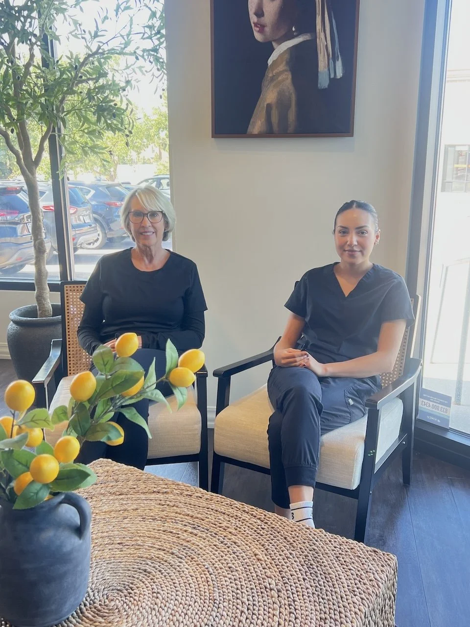 Two women in black scrubs sit in armchairs in a bright waiting area with a lemon plant on the table and a framed portrait behind them.