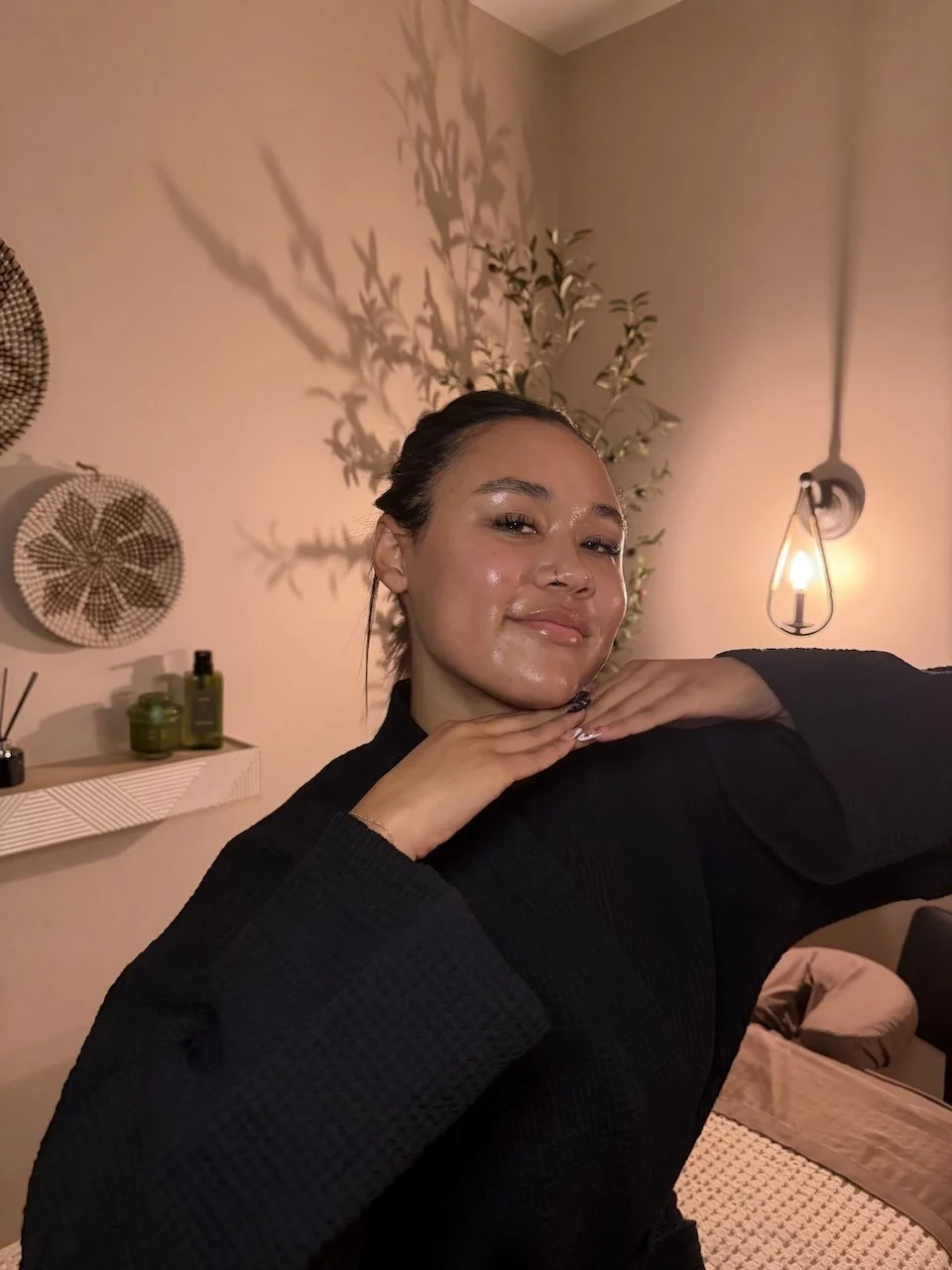 A person in a black robe poses with hands under chin, smiling, in a warmly lit room with skincare products and a wall lamp in the background.