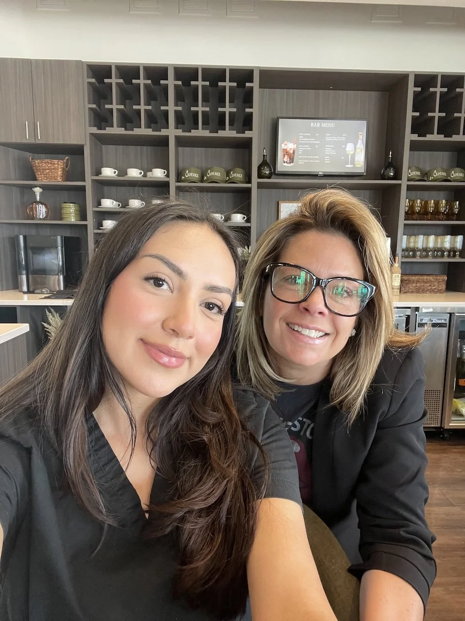 Two women take a close selfie inside a modern salon or spa, smiling at the camera, with shelves of cups, bottles, and a bar menu visible in the background.
