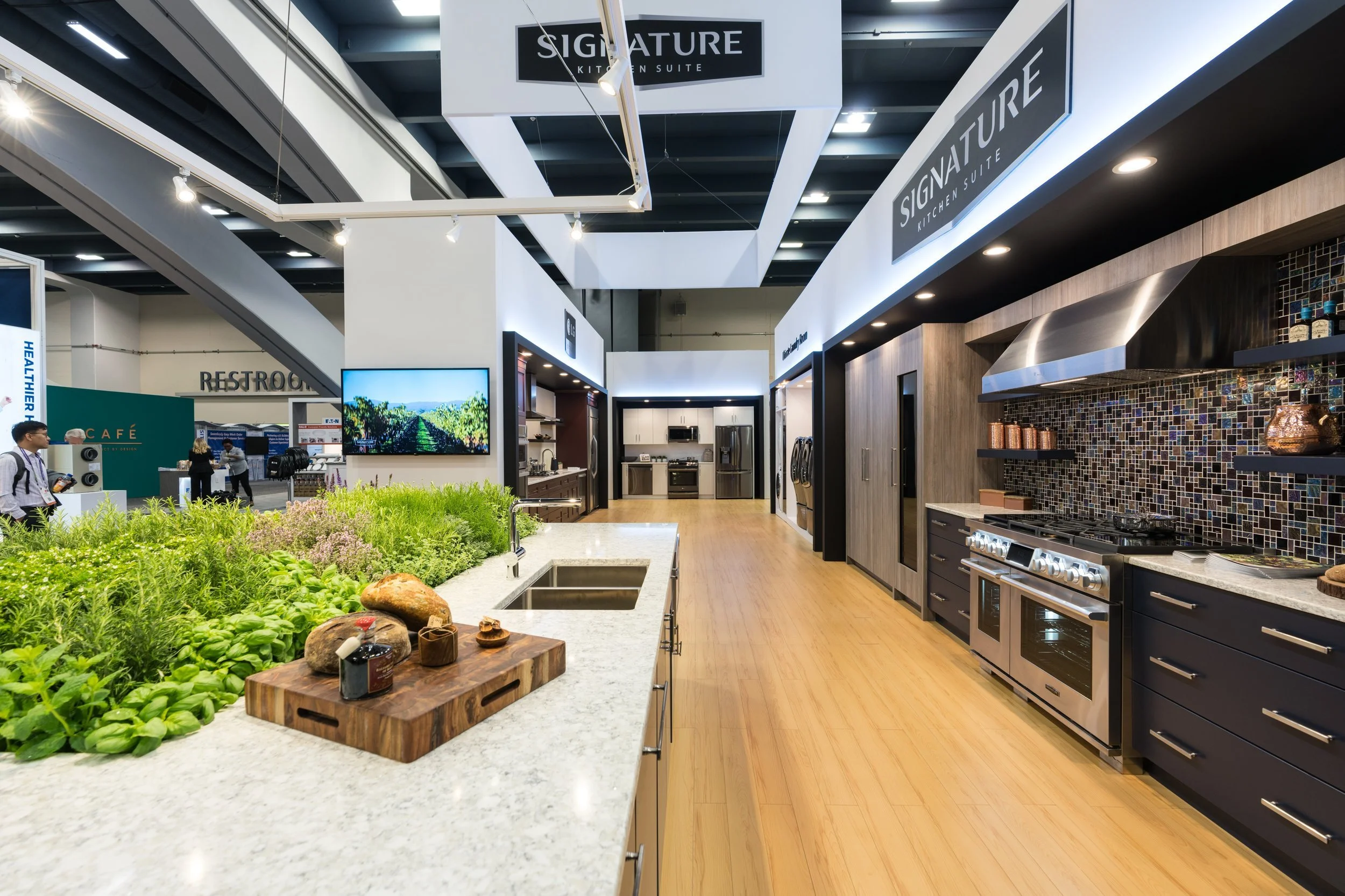 An LG exhibition kitchen display with modern appliances, dark cabinets, a mosaic backsplash, and a wooden floor, for a trade show. Created by MEGA Exhibit.