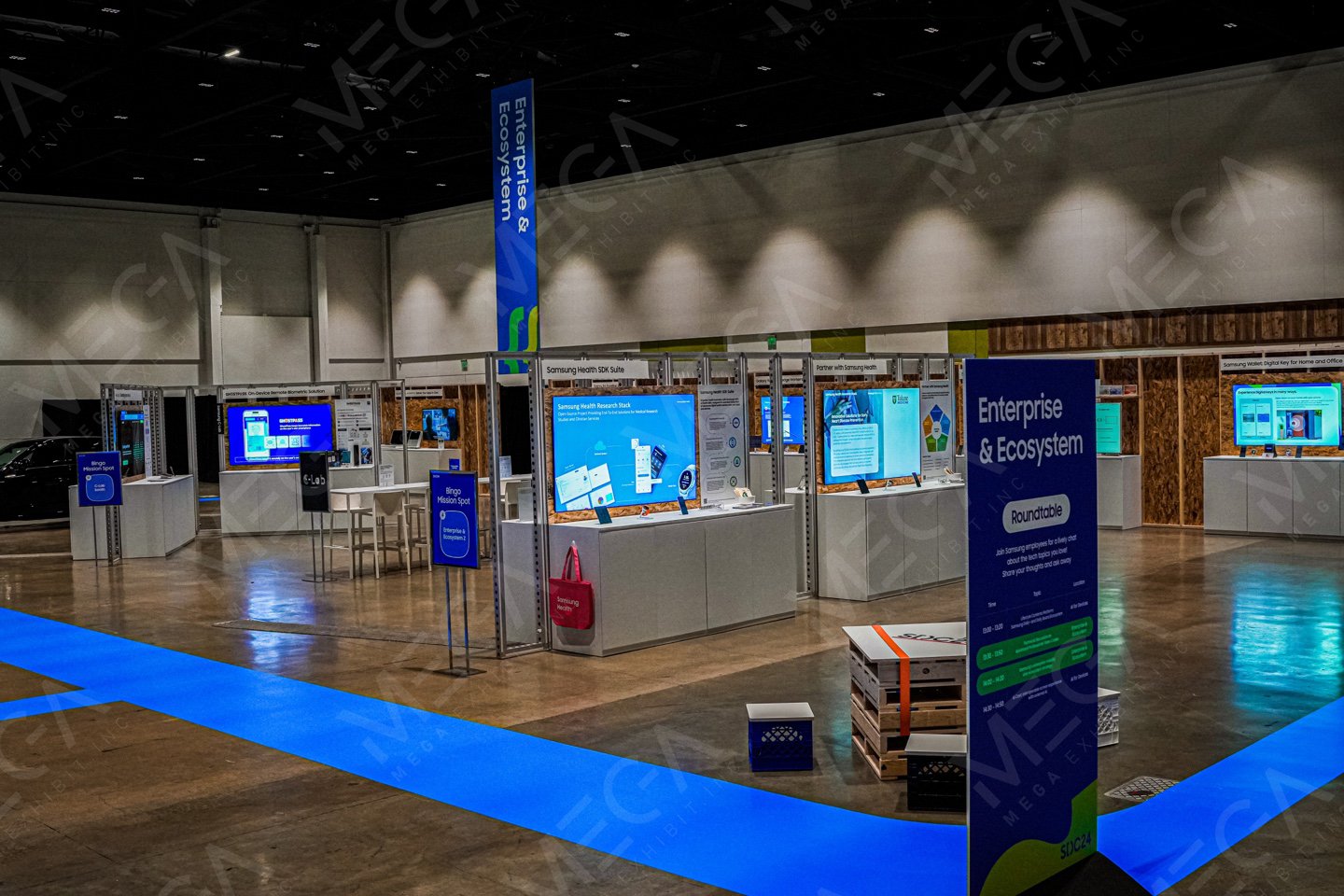 Exhibition booth for Samsung's SDC 2024 labeled 'Enterprise & Ecosystem' with multiple screens displaying digital content, standing desks, and signs, set up in a large indoor event space with sustainable materials.