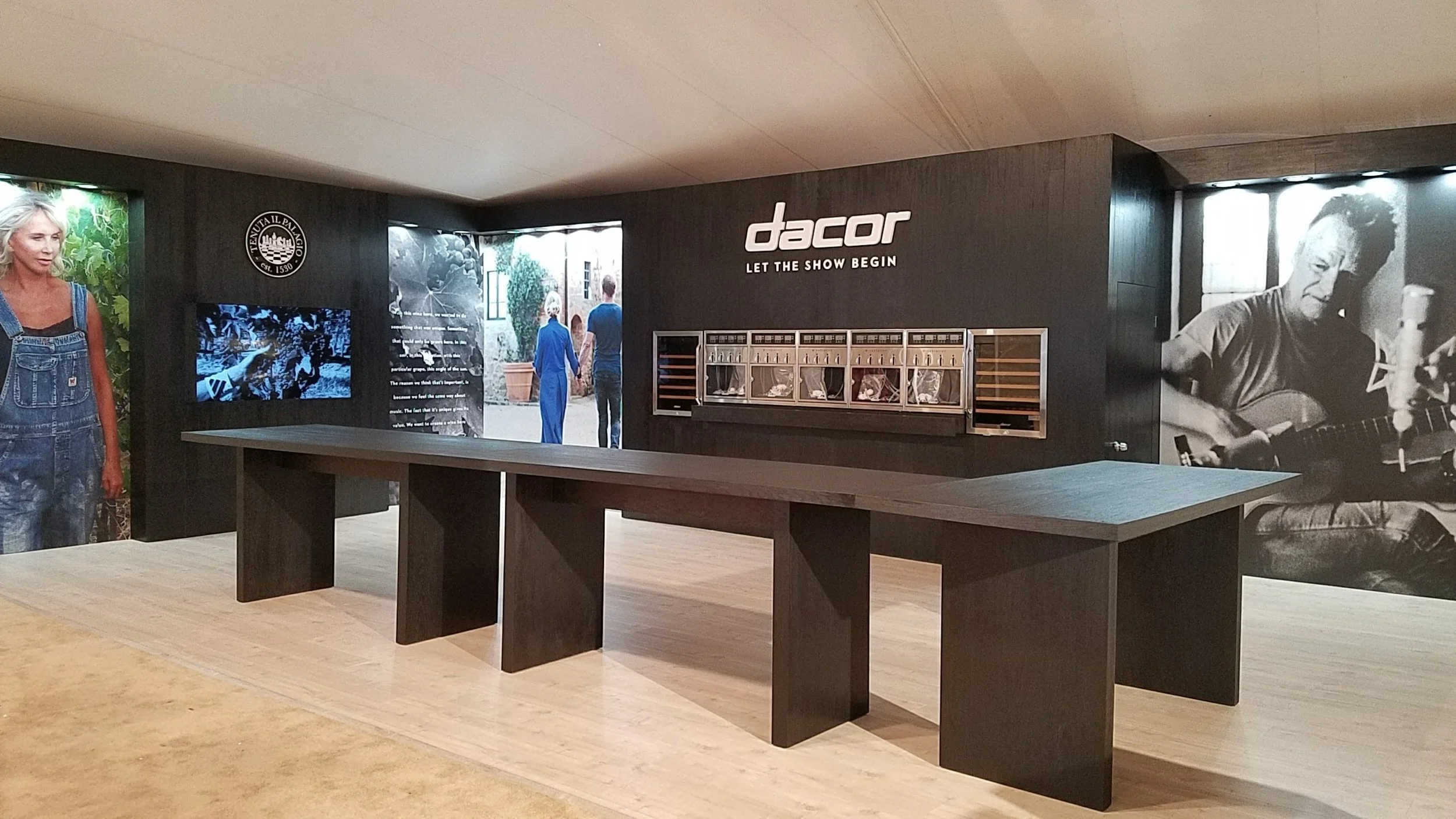 Interior of a showroom with black walls, large images and videos, and a table in the center, promoting the brand 'dacor' with the slogan 'Let the Show Begin'. Created by MEGA Exhibit.