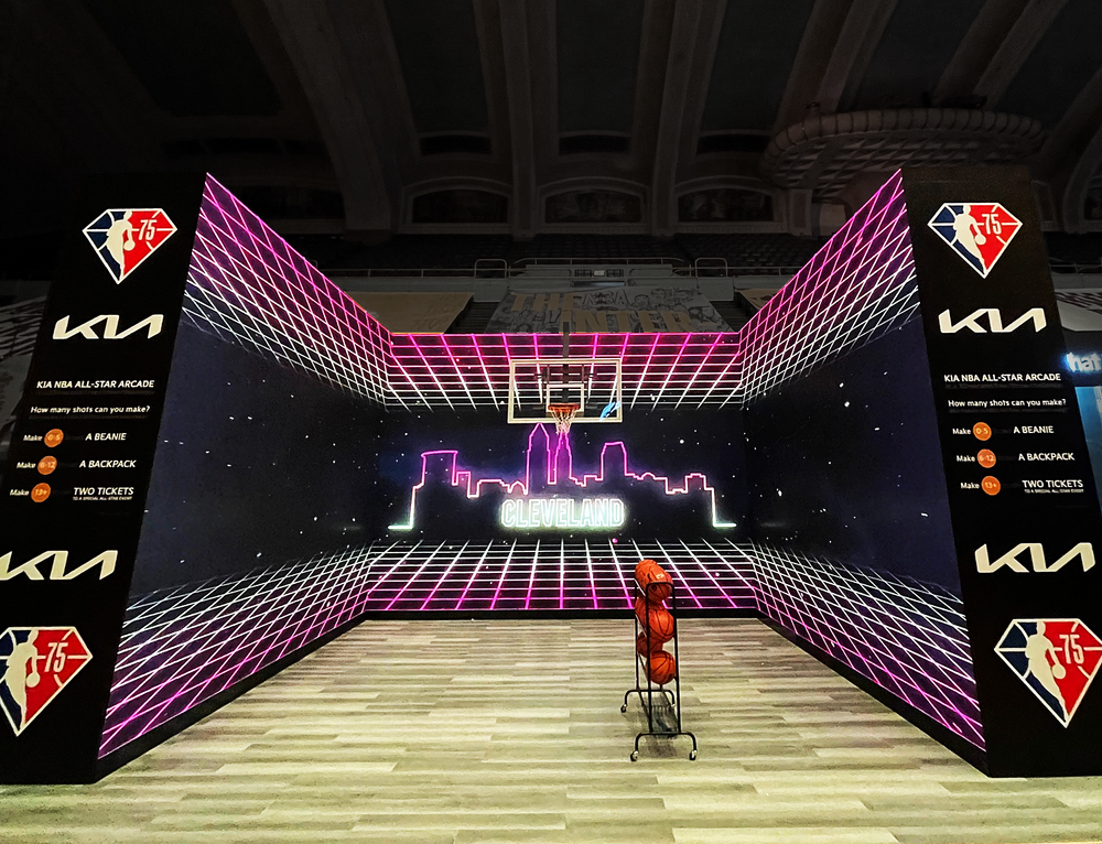 Indoor basketball court exhibit booth with a neon Cleveland skyline and cityscape on a digital screen, pink grid pattern on the walls, basketballs on a cart in front, and NBA 75th anniversary logos on side panels for Kia.