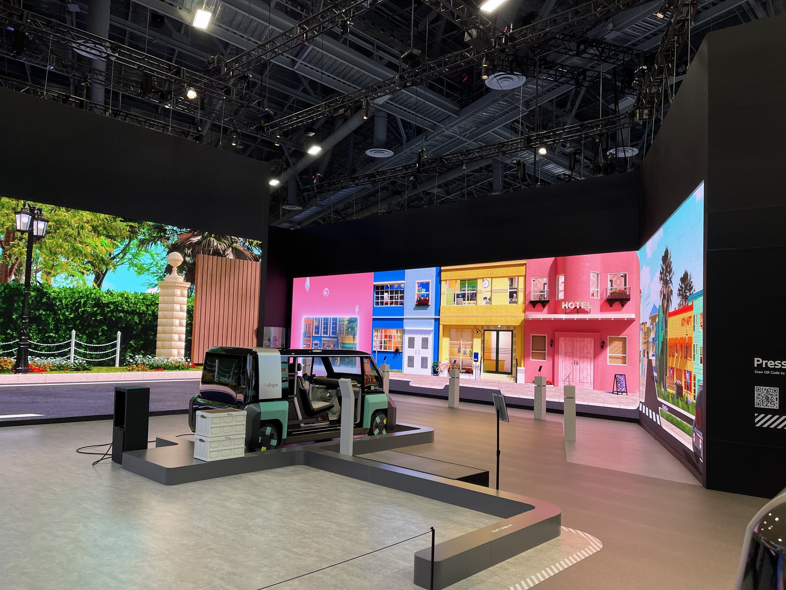 Exhibit at SDC 2024 by Samsung. It features a small, model electric vehicle with LED displays in a large indoor exhibition space with digital screens showing colorful cityscape scenes. Created by MEGA Exhibit.