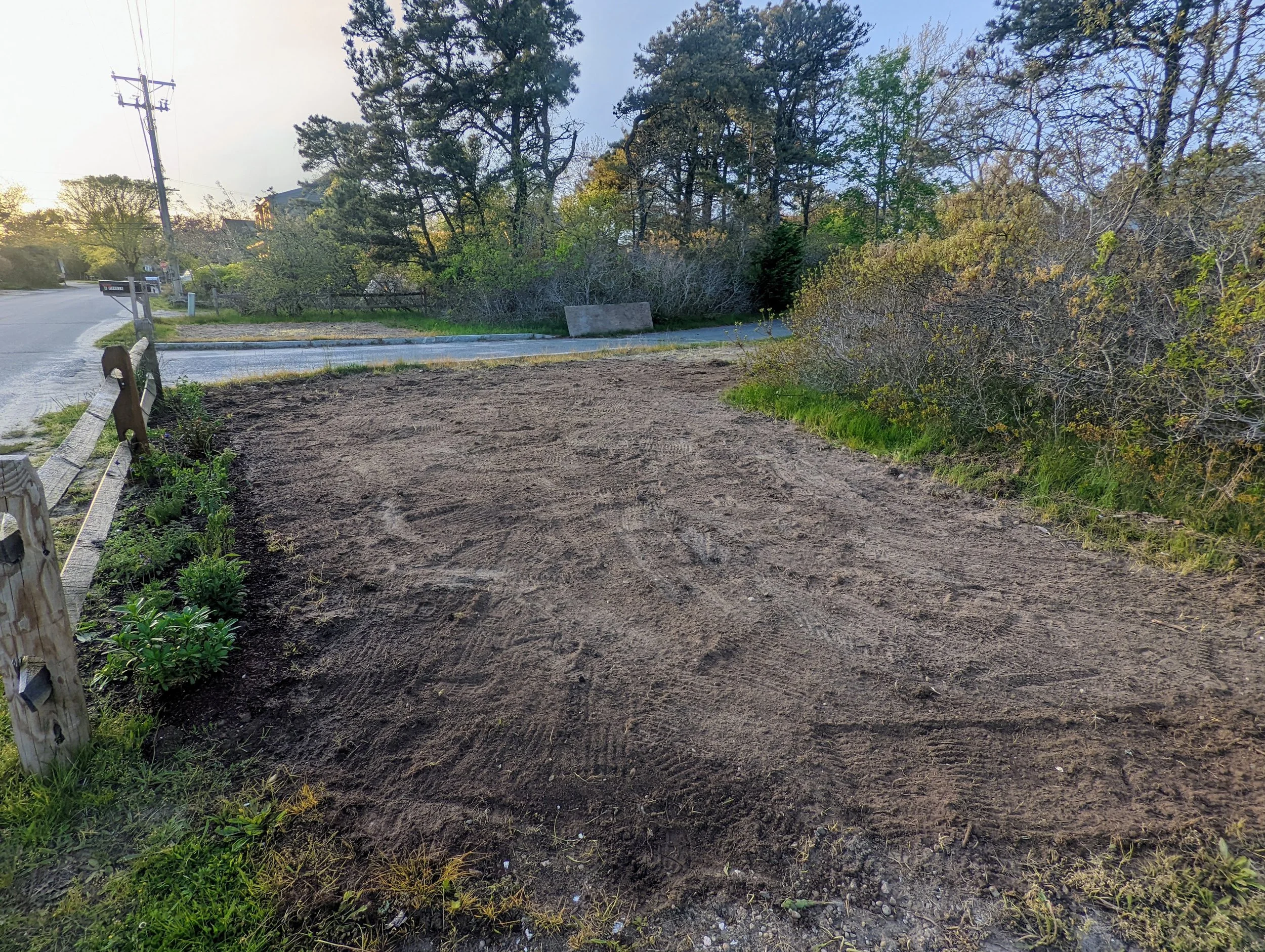 Empty dirt lot beside a street with trees and bushes, a park bench, and a wooden fence, during daytime.