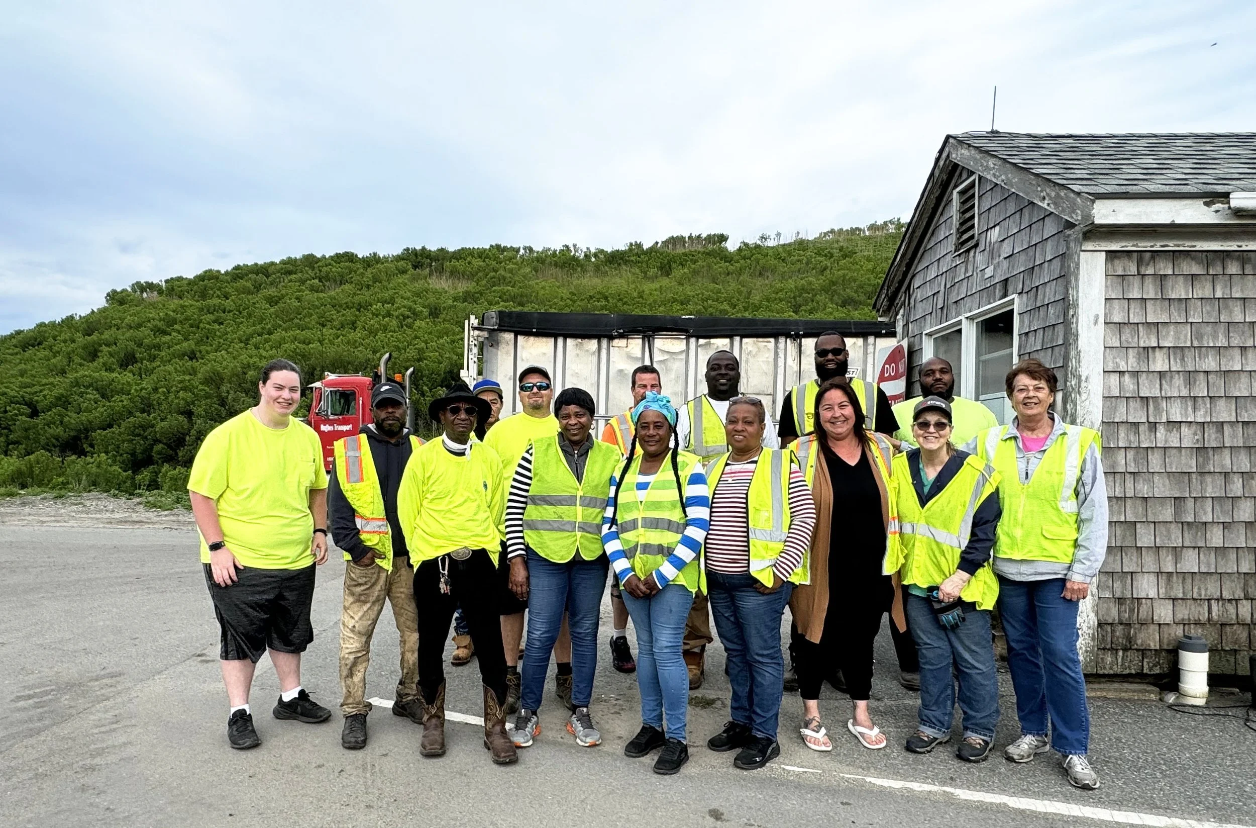 Group of people wearing high-visibility vests standing outdoors in front of a building and a truck with greenery in the background.