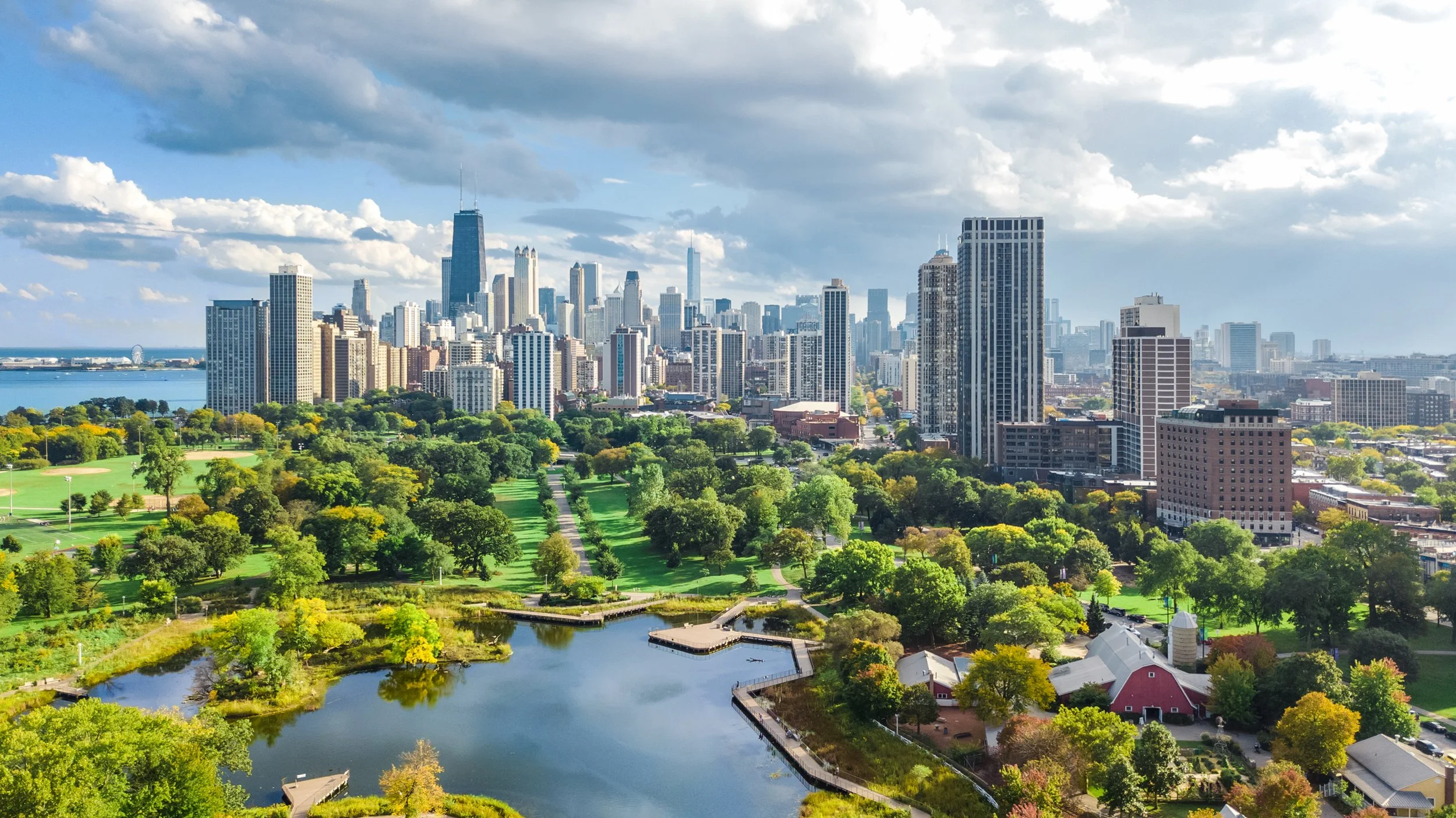 A city skyline with numerous tall buildings and skyscrapers, a large urban park with green trees, a pond, and walking paths, under a partly cloudy sky.