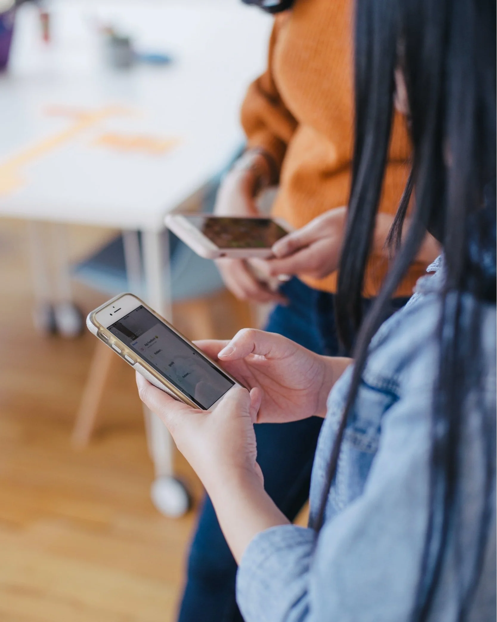Two people are holding smartphones, with one person wearing a denim jacket and another wearing an orange sweater, in an indoor setting.
