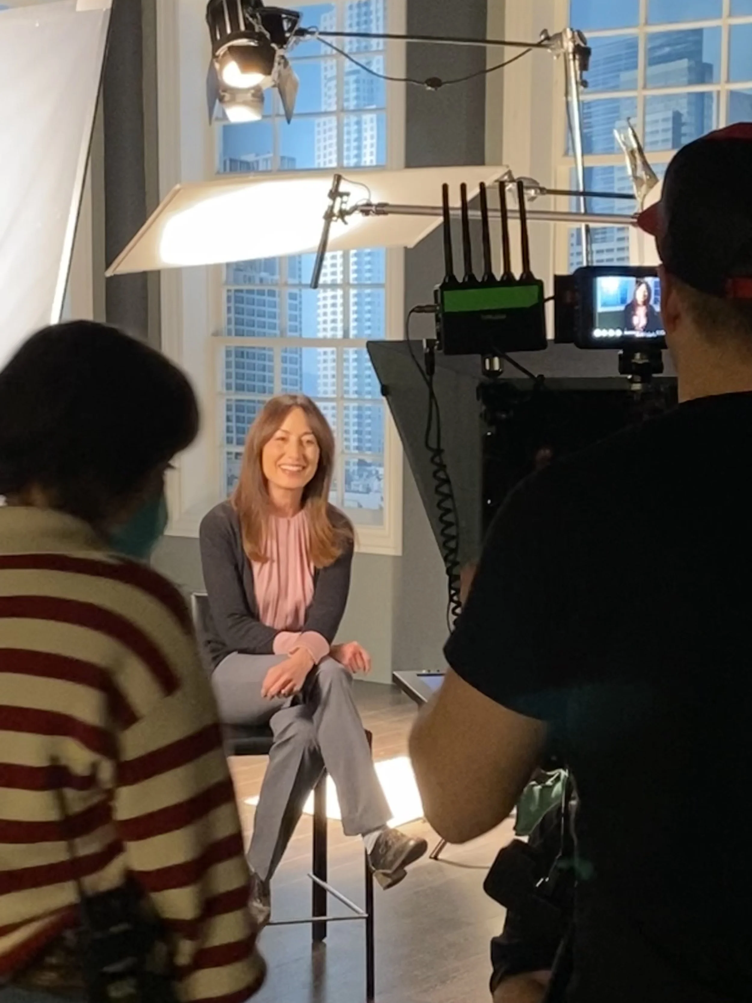 A woman with long reddish-brown hair is sitting on a stool, smiling during a video shoot. She is wearing a pink shirt, gray pants, and a dark blazer. There are studio lights set up around her, with a camera recording her in an indoor setting with large windows and a cityscape view in the background.