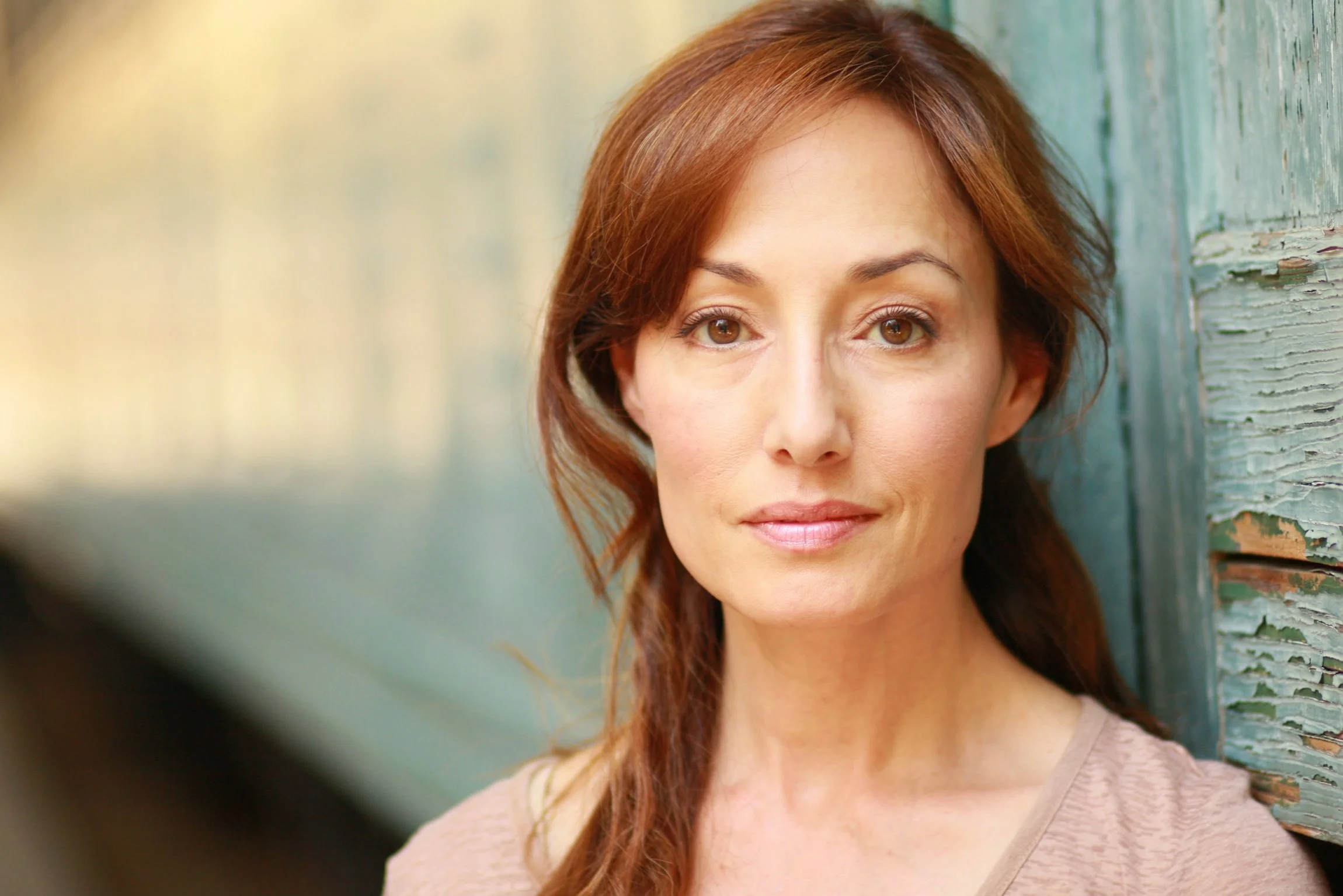 Close-up of a woman with light brown hair, standing against a weathered turquoise wooden wall, looking at the camera.