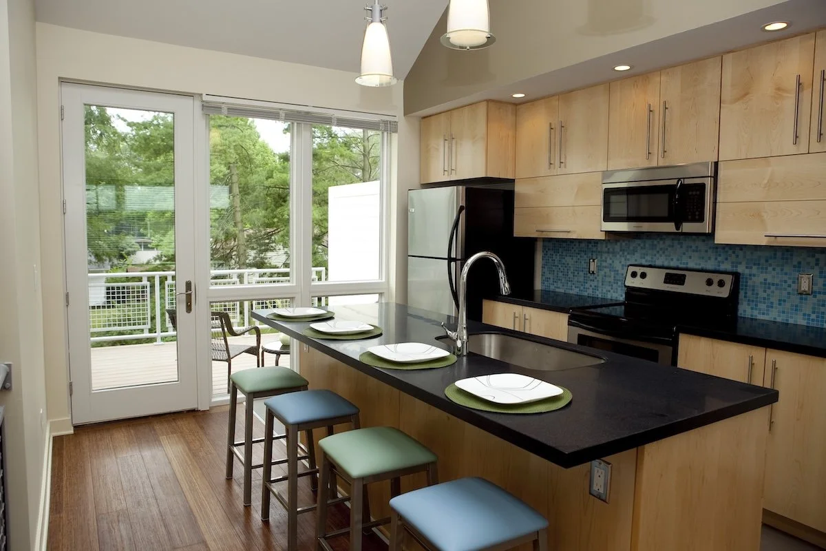 Modern kitchen with light wood cabinets, black countertops, a teal tile backsplash, and stainless steel appliances. There's a kitchen island with four stools and pendant lighting above. Large windows provide natural light, and a potted plant is seen 