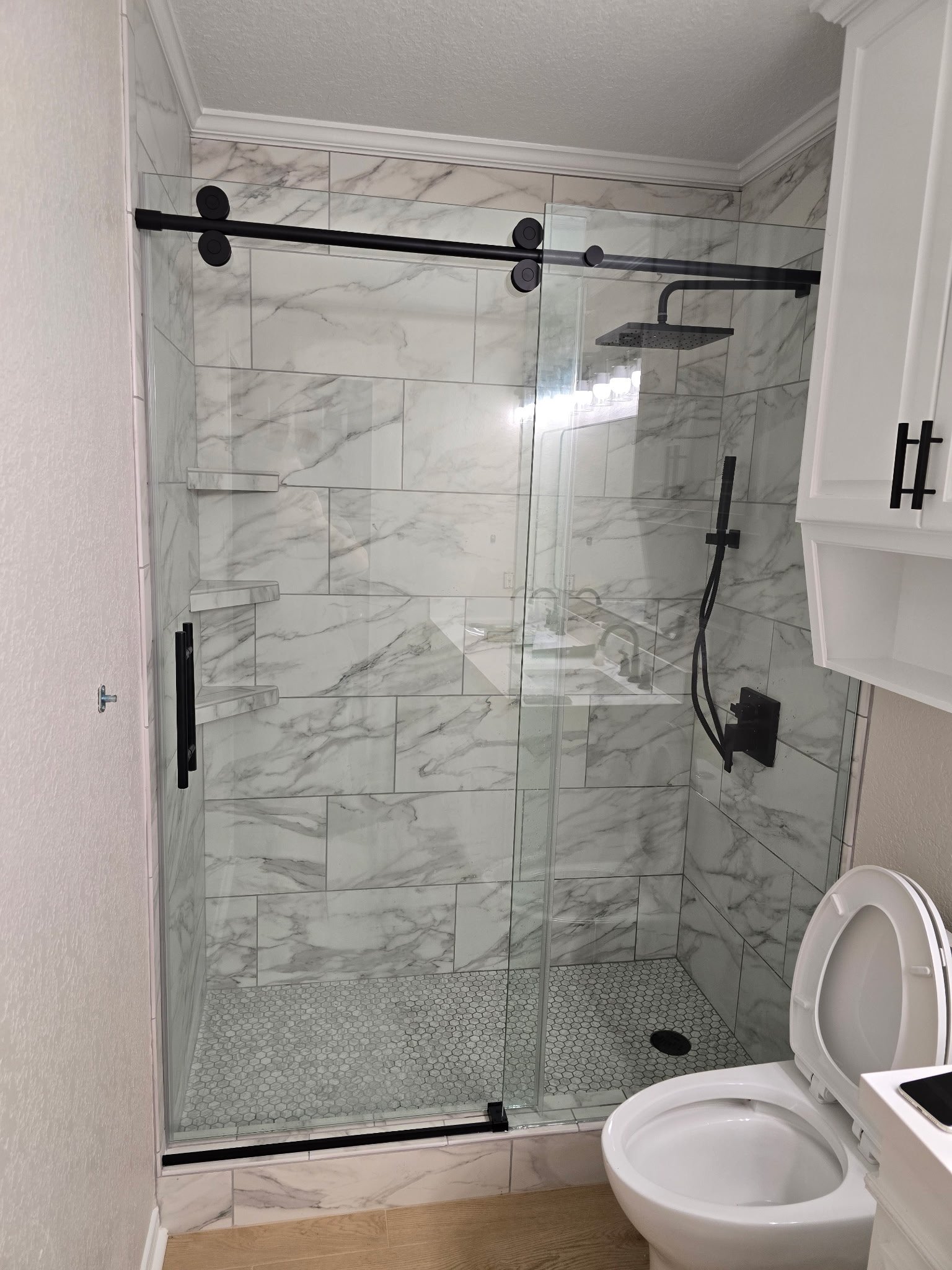 Bathroom with marble-tiled shower enclosure, black fixtures, glass door, and a toilet, with a wooden floor and white walls.