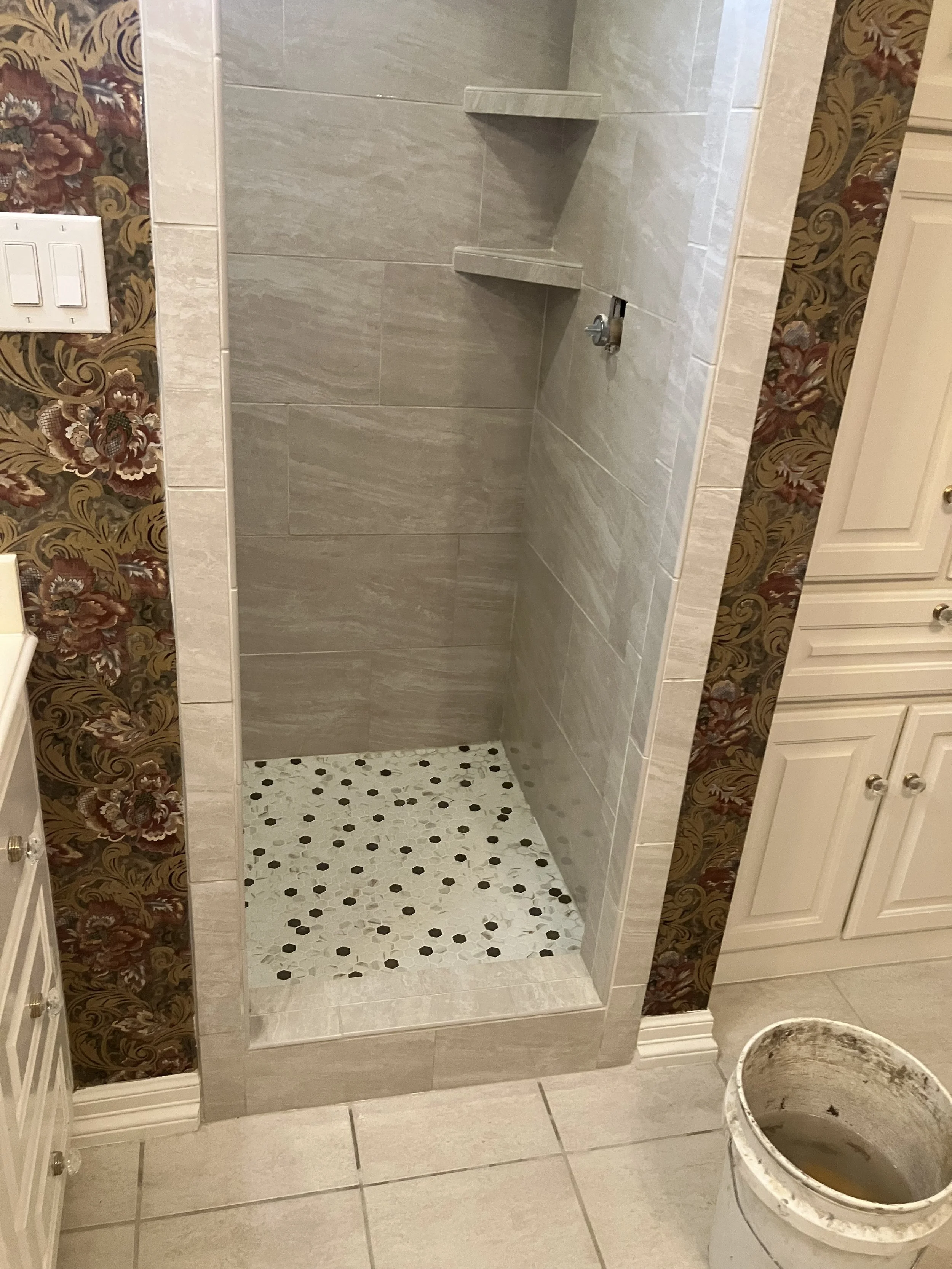 Empty shower stall with beige tiles, corner shelves, floral wallpaper on the walls, and a bucket on the tiled floor outside the shower.