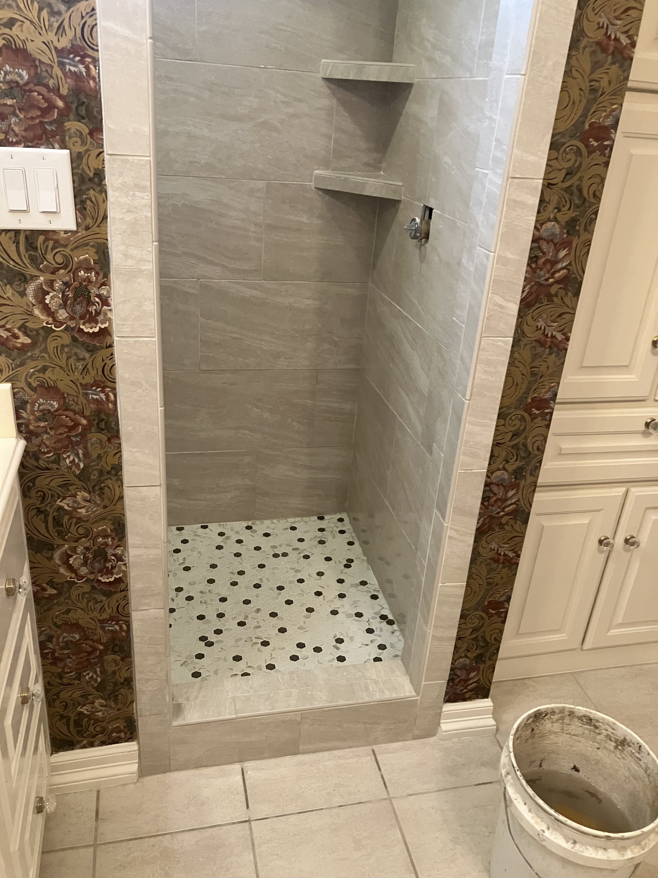 Empty shower stall with beige tiles, corner shelves, floral wallpaper on the walls, and a bucket on the tiled floor outside the shower.