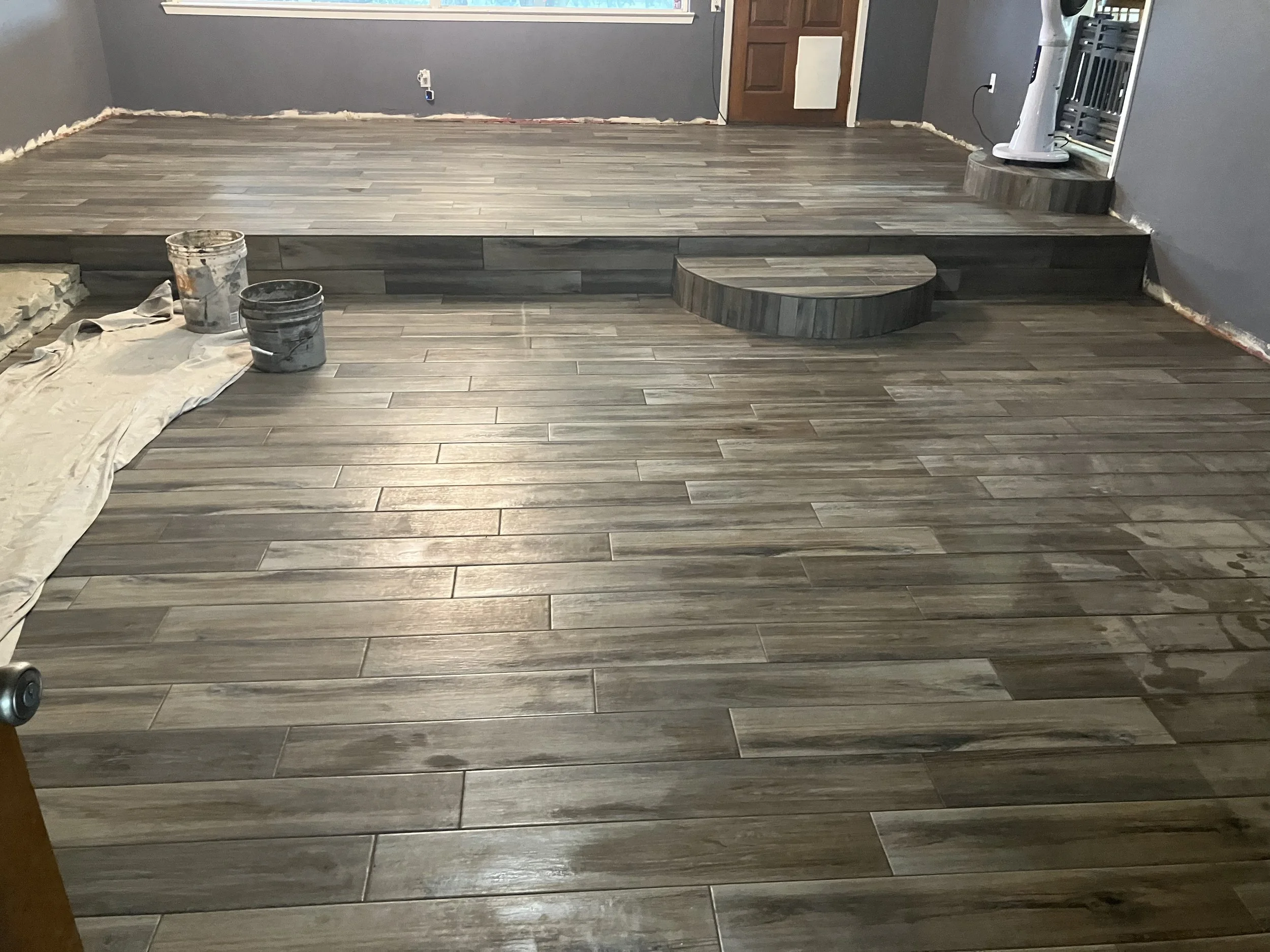 Interior of a room under renovation with newly installed wood-look tile flooring. There are two buckets of grout or paint on a cloth on the left side, and a small semi-circular step leading up to a raised area at the back of the room. A fan and some construction tools are visible near the right wall. The walls are painted gray, and there is a window, door, and electrical outlet visible.