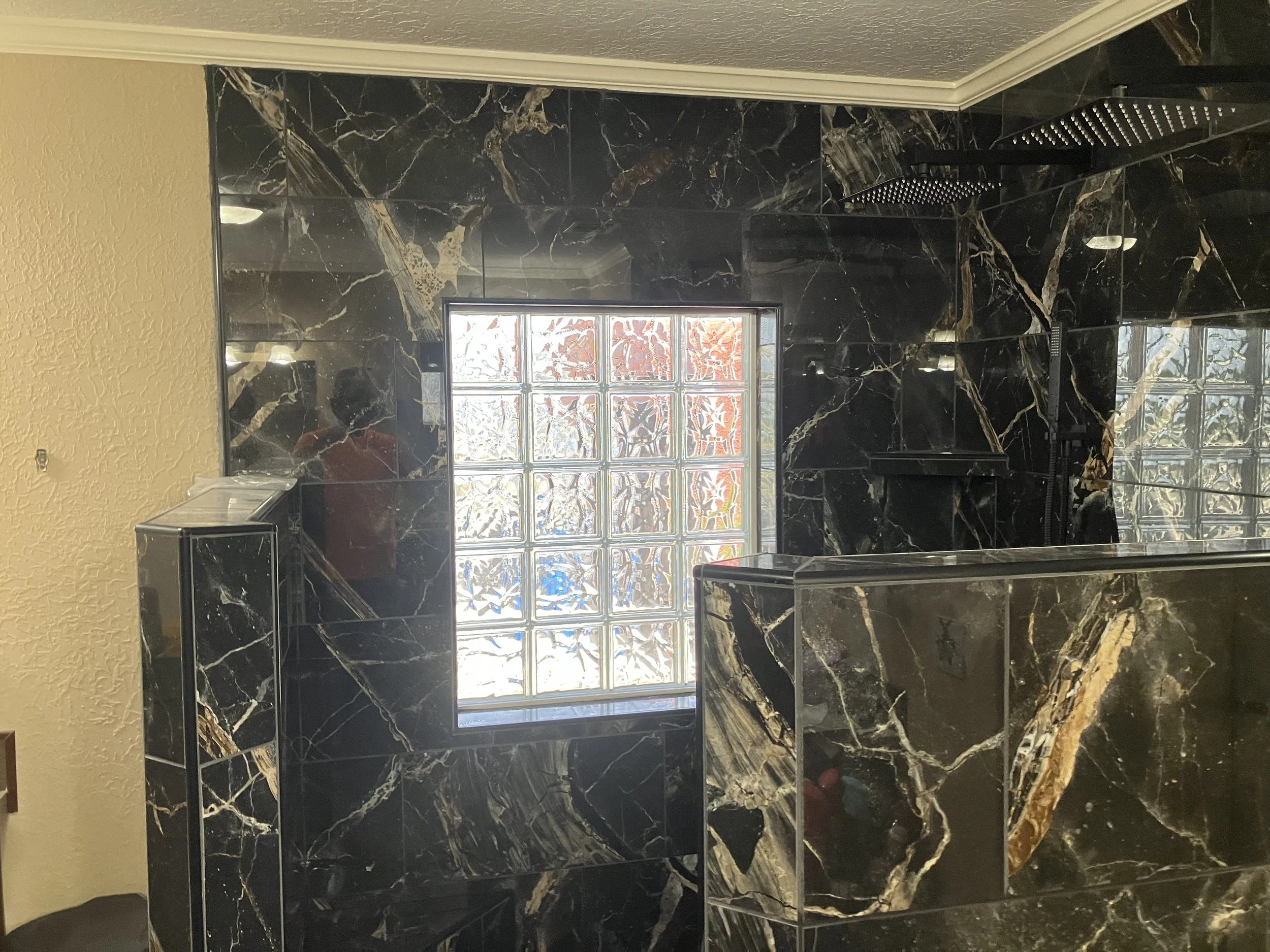 A bathroom with black marble tiles featuring white and gold veining, a small window with decorative glass blocks, and a black rain showerhead.