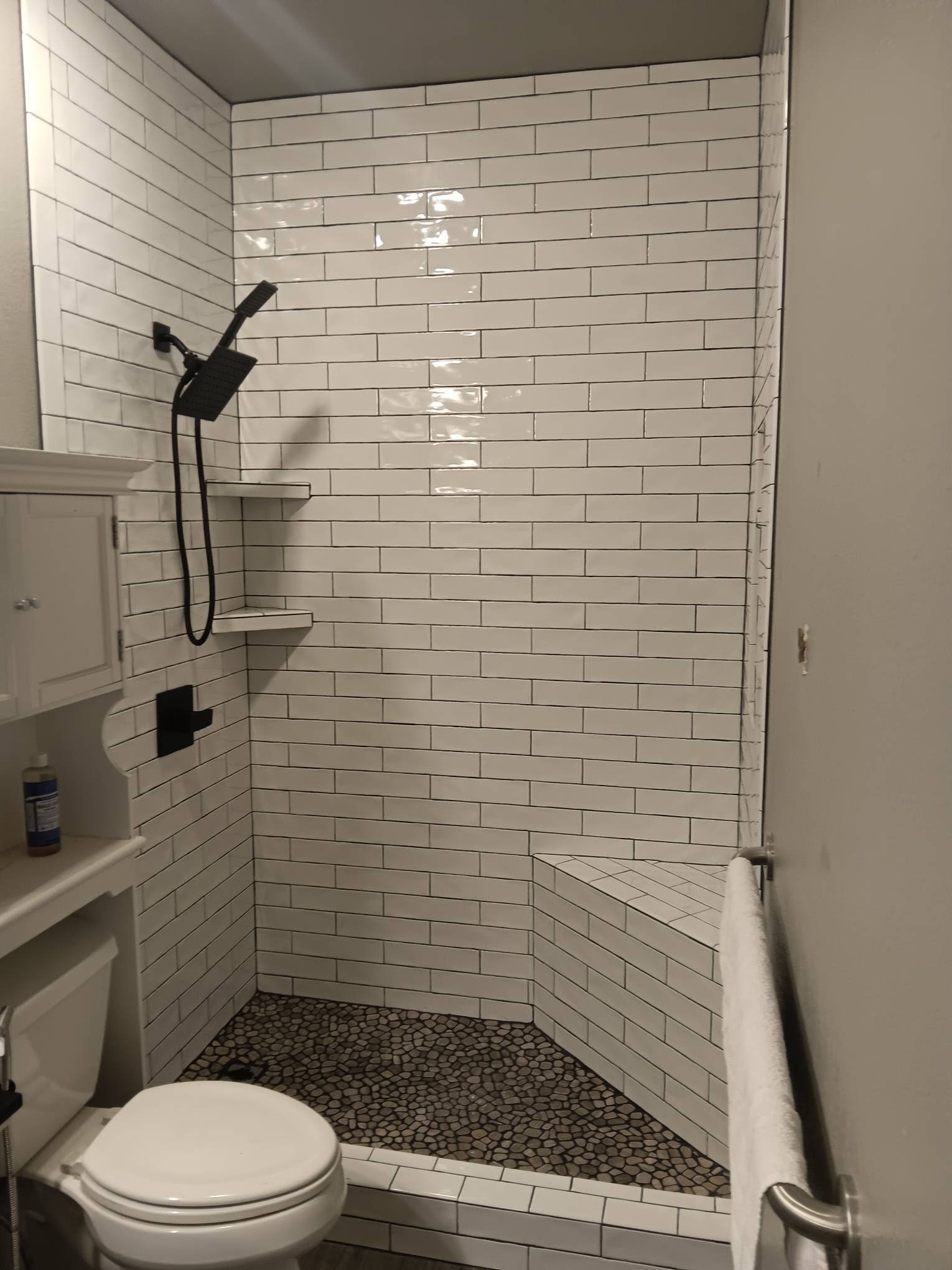 Bathroom shower with white subway tiles, a black showerhead and controls, built-in benches, pebble floor, and a wall with a towel rack.