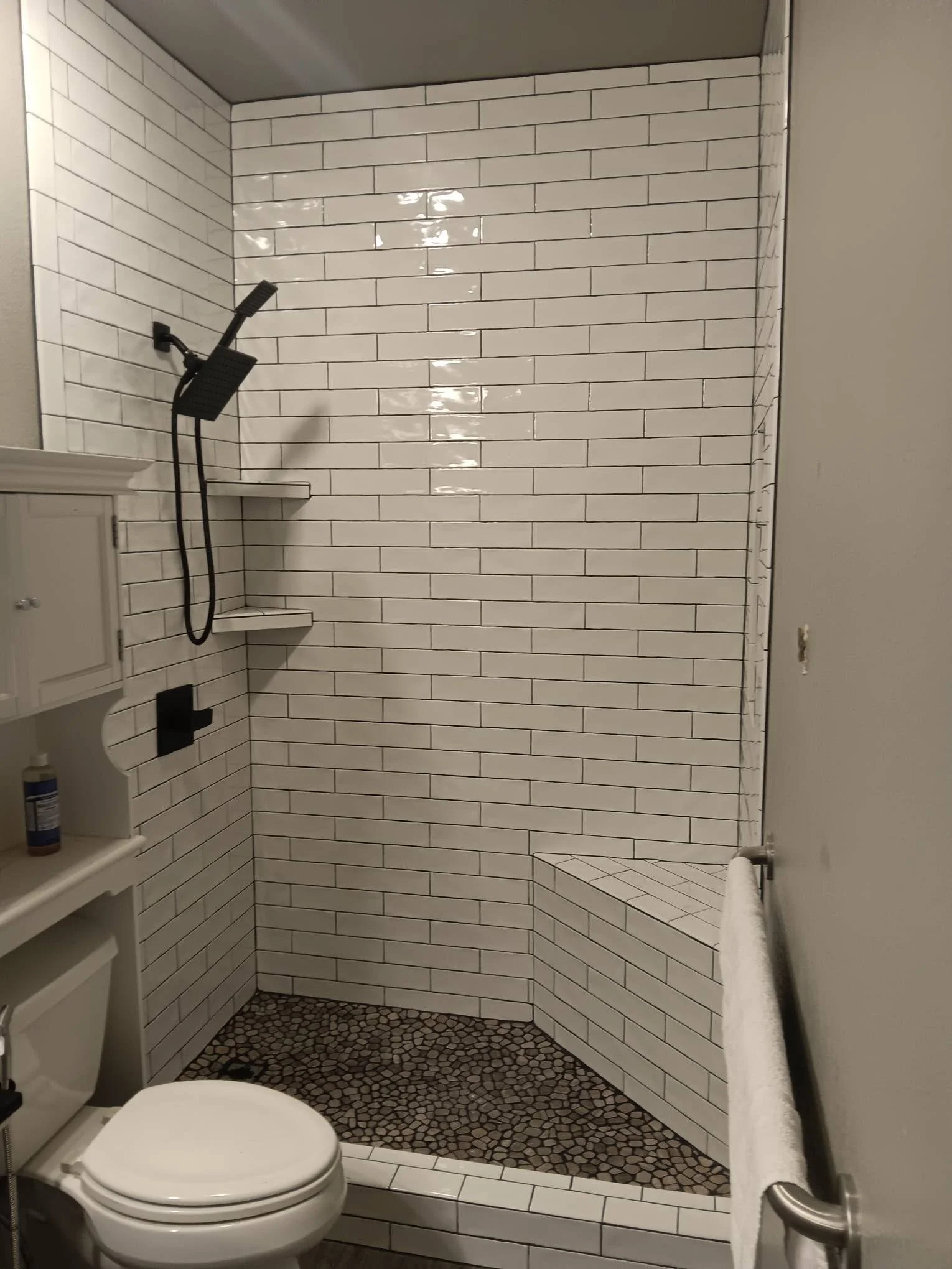 Bathroom shower with white subway tiles, a black showerhead and controls, built-in benches, pebble floor, and a wall with a towel rack.