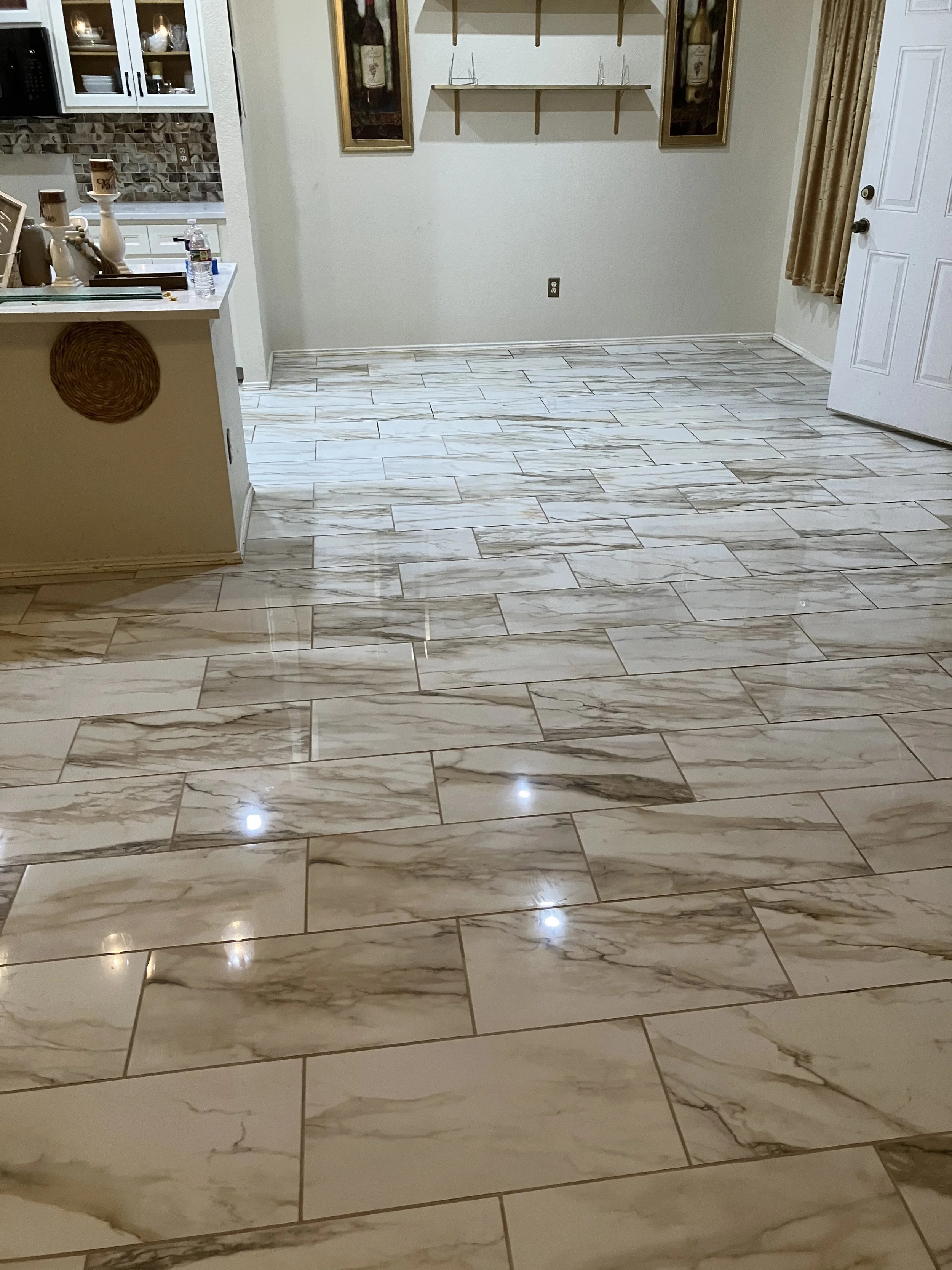 Living room with a beige marble tile floor, part of a white kitchen counter with various items, wall art and shelves, a white door, and a window with gold curtains.
