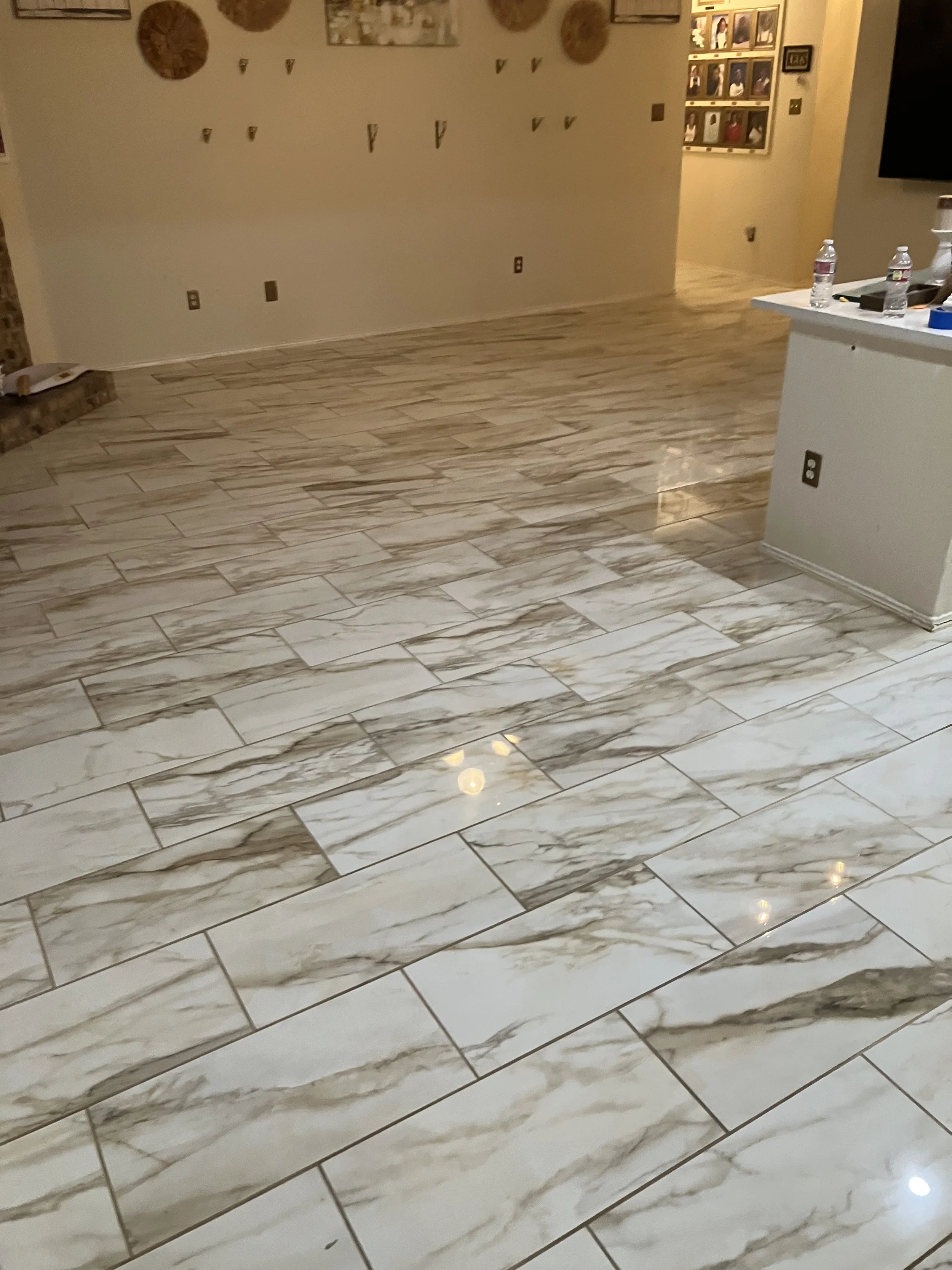 Photo of a room with tru marmi 12x24 tile flooring, light-colored walls decorated with baskets and small wall hooks, a small section of brick fireplace, and a white counter with water bottles and other items.