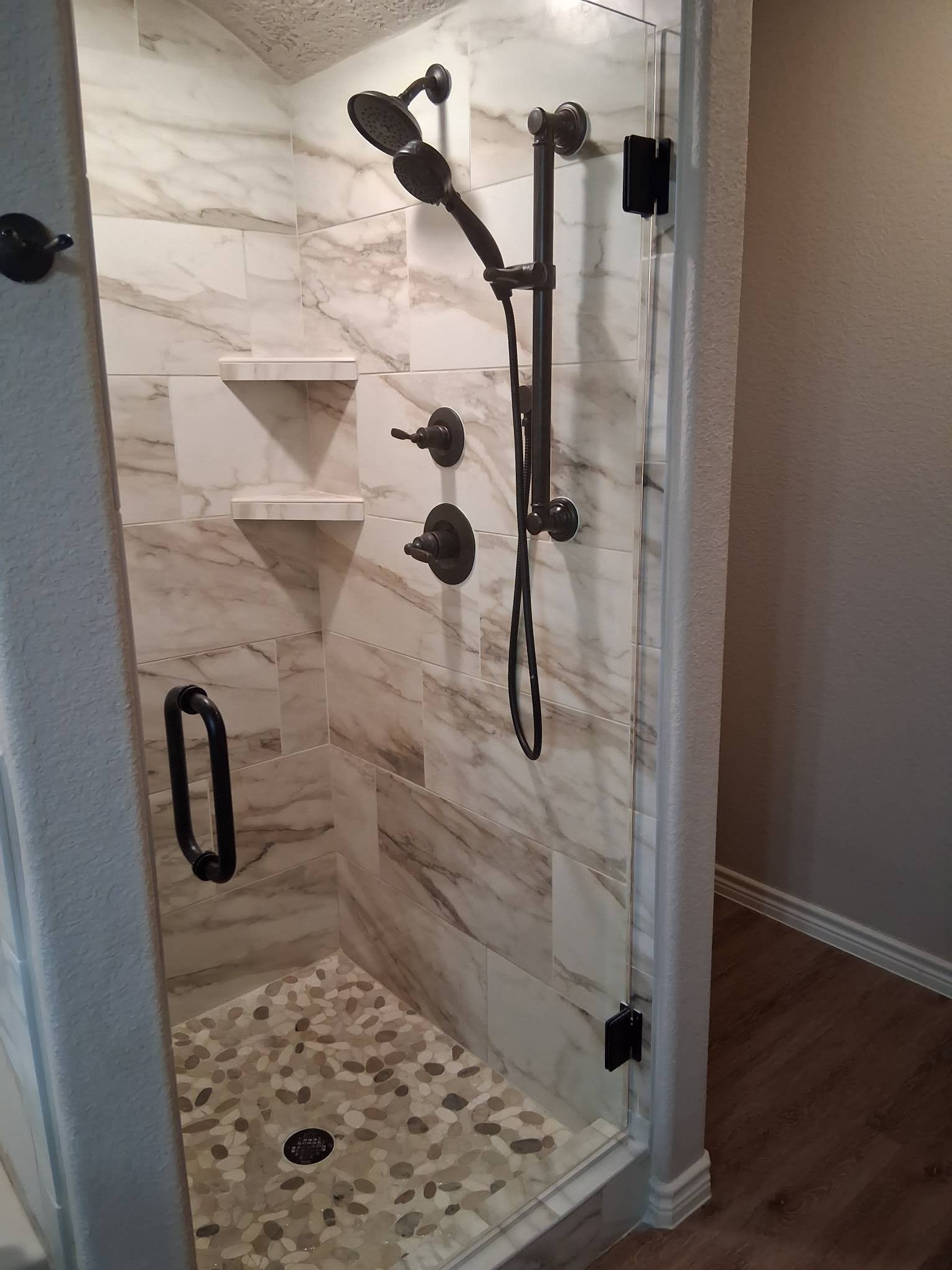 A walk-in shower with marble tile walls, a pebble tile floor, and black fixtures, including a showerhead, handheld shower, multiple knobs, and a glass door with a black handle.