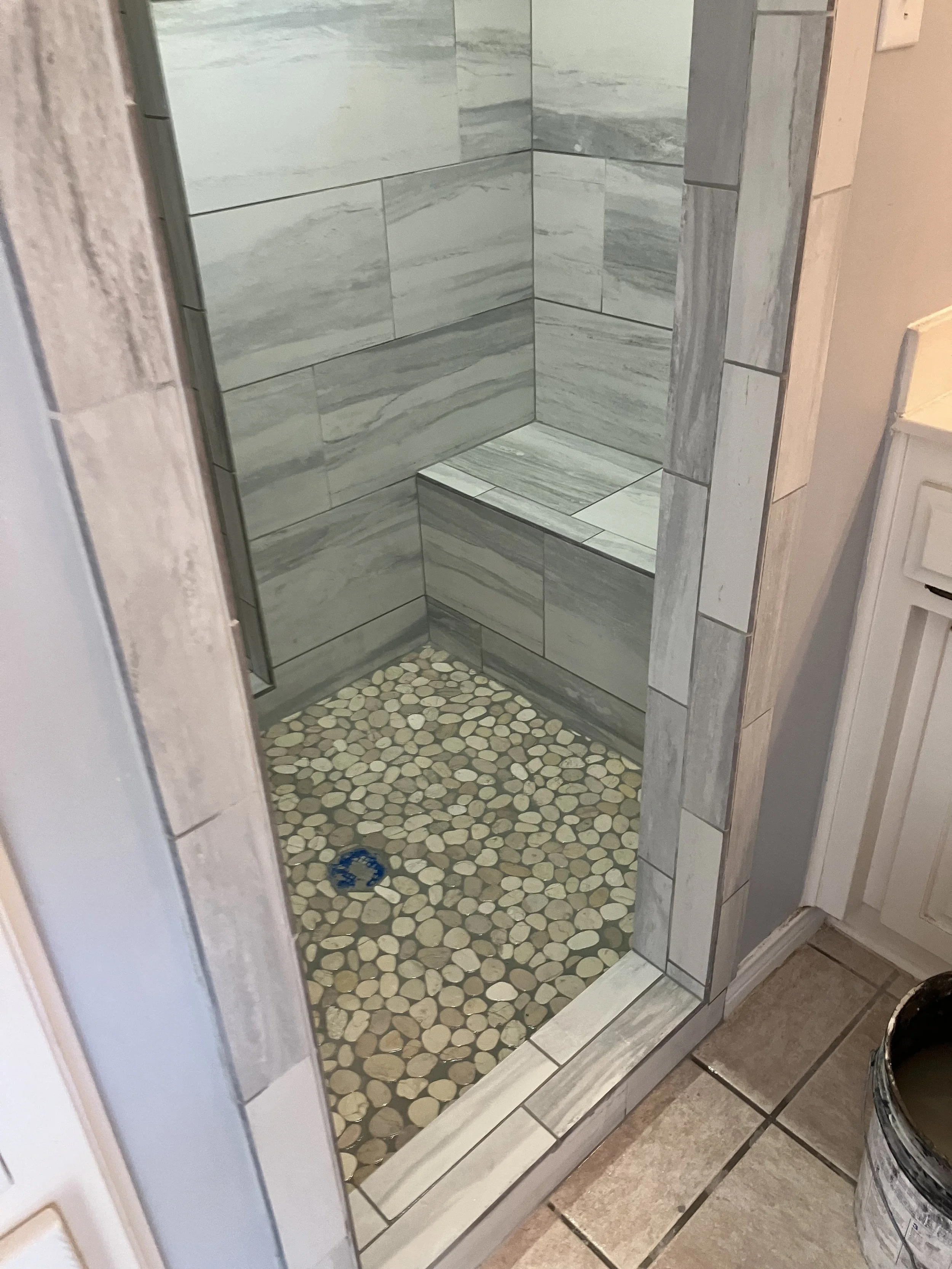 A small shower with gray marble tiles on the walls and pebble stones on the floor, with a mirror reflecting part of the bathroom.