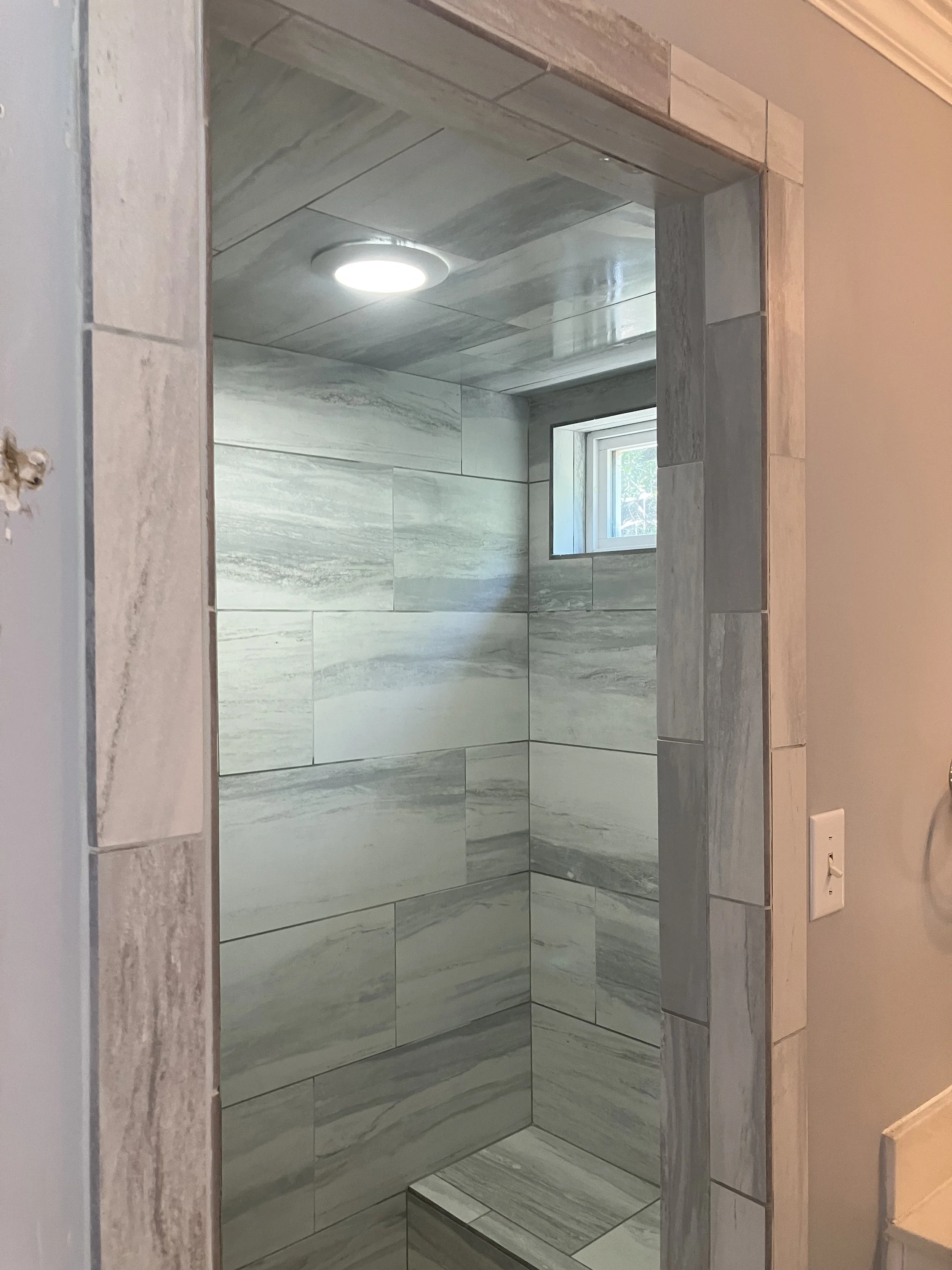 Inside a newly tiled bathroom shower with gray marble tiles, a small window, and a recessed ceiling light.