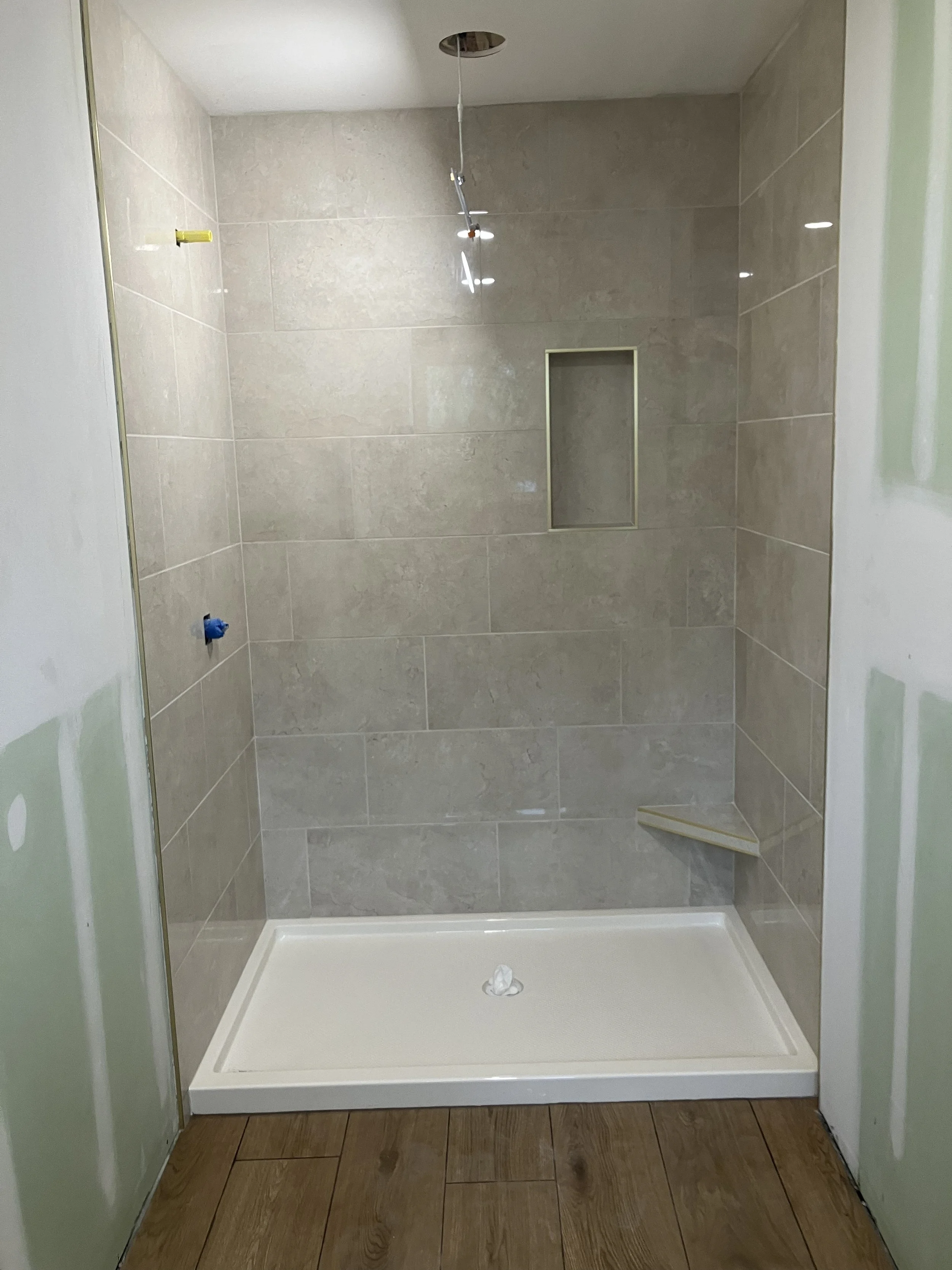 Unfinished shower with beige tiled walls, a small recessed shelf, a showerhead hanging from the ceiling, and brown tile flooring outside the shower.