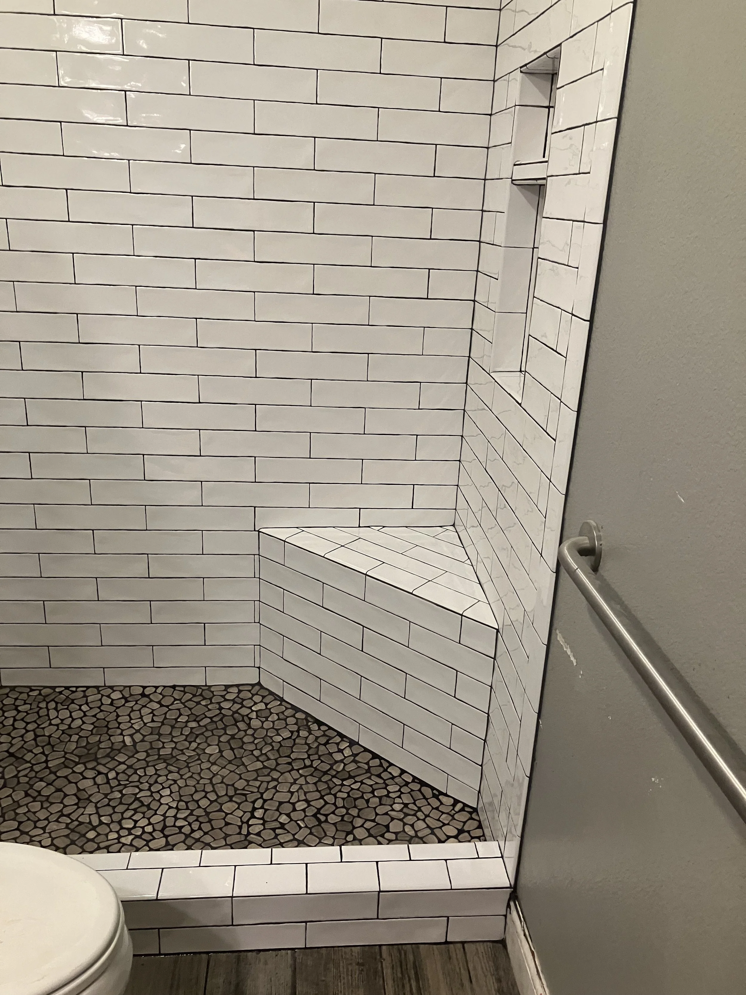 Close-up of a tiled shower with a built-in seat, white subway tiles on the walls, pebble tiles on the floor, and a niche for toiletries, with a gray wall and towel bar outside the shower.