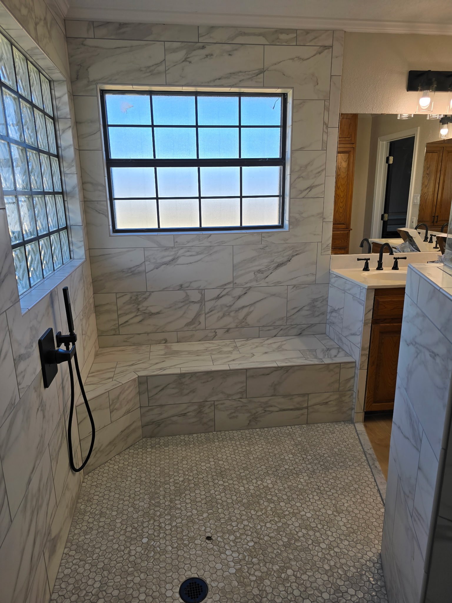 A modern bathroom shower area with white marble tiles, a frosted window, and a separate vanity with wooden cabinets and black fixtures.