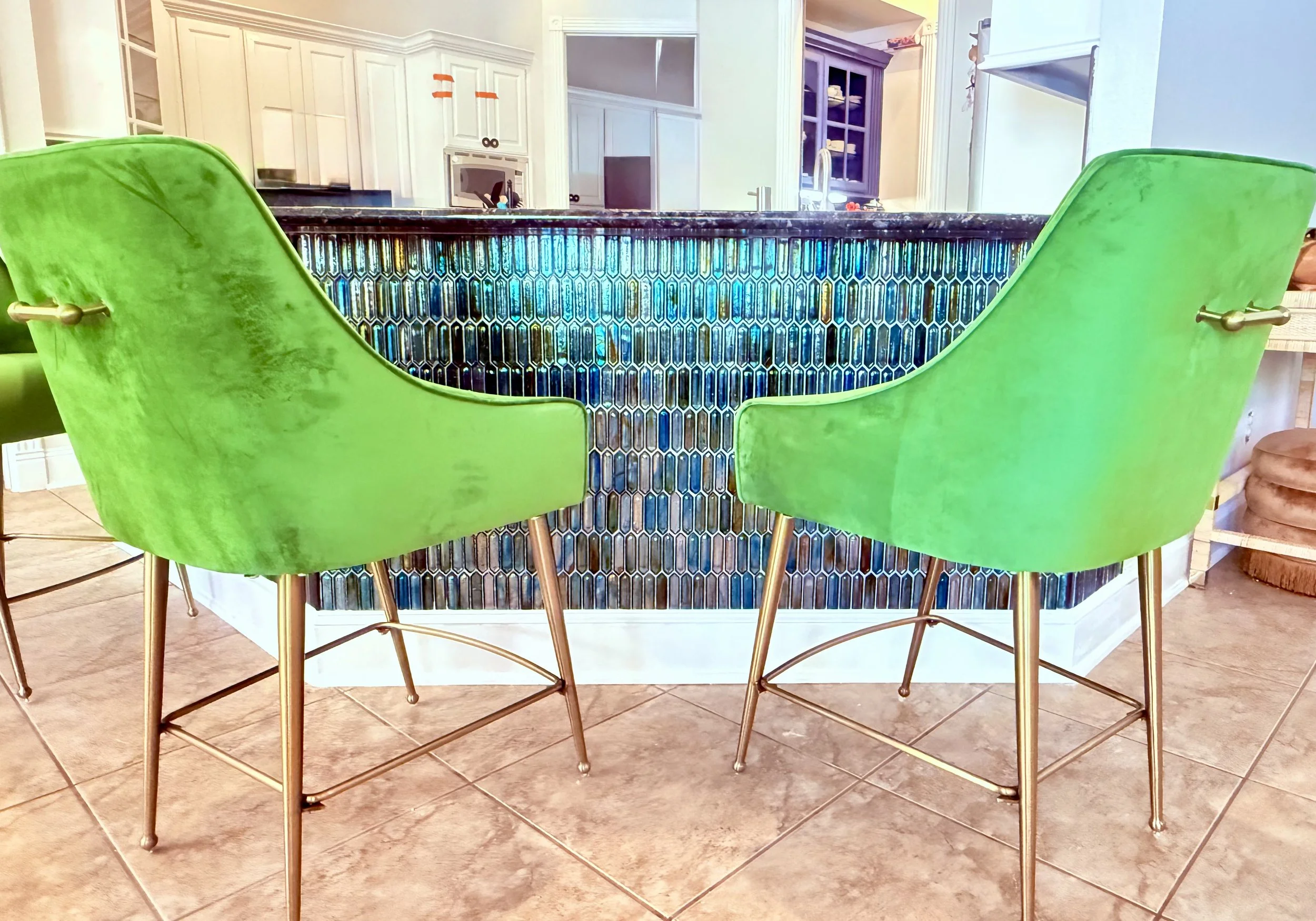 Bright green velvet chairs around a high counter with colorful mosaic tiles, in a kitchen with white cabinets and black countertops.