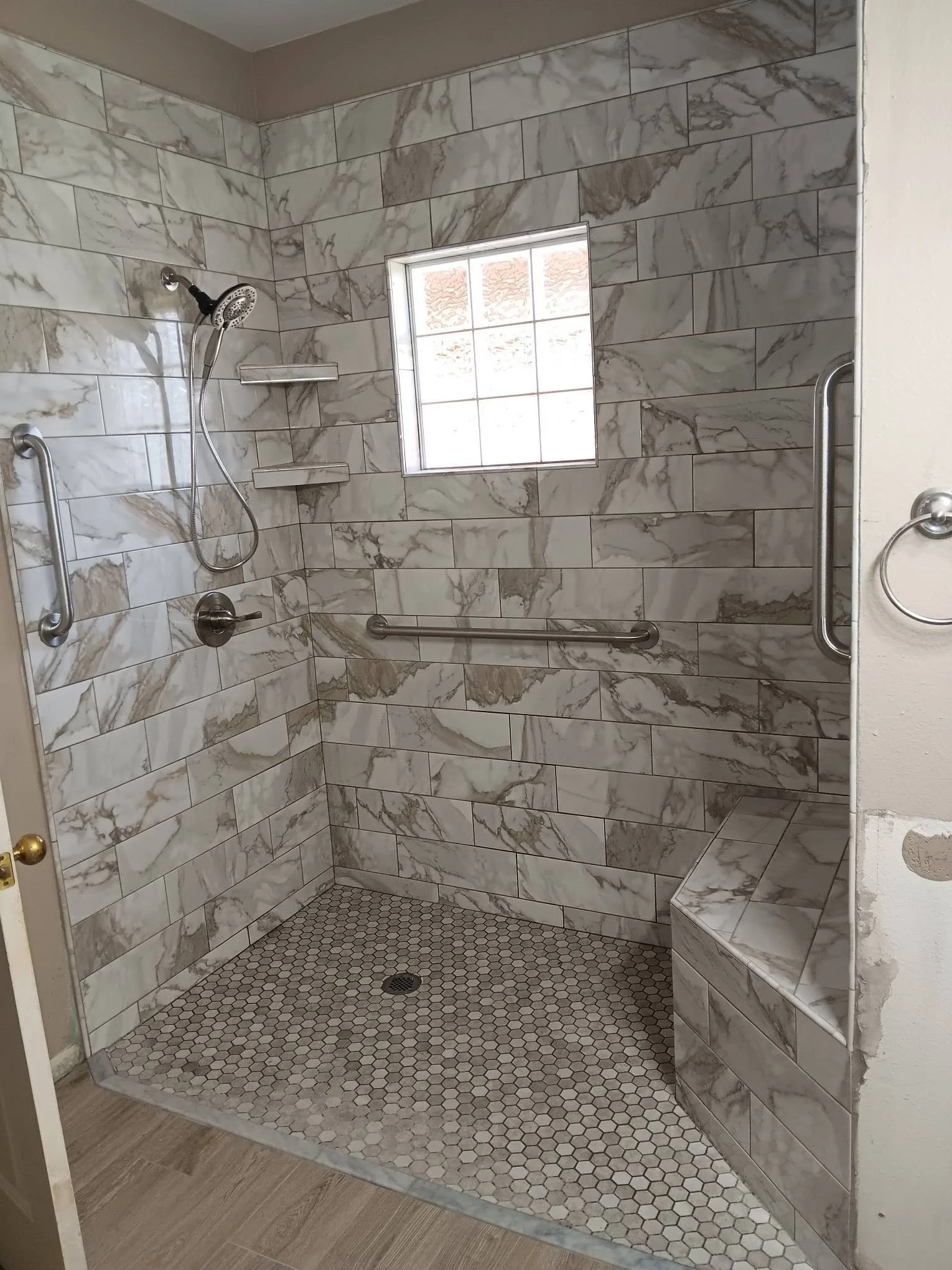 A walk-in shower with marble wall tiles, a small window, a handheld showerhead, a grab bar, and built-in seating, with a wooden floor outside the shower.