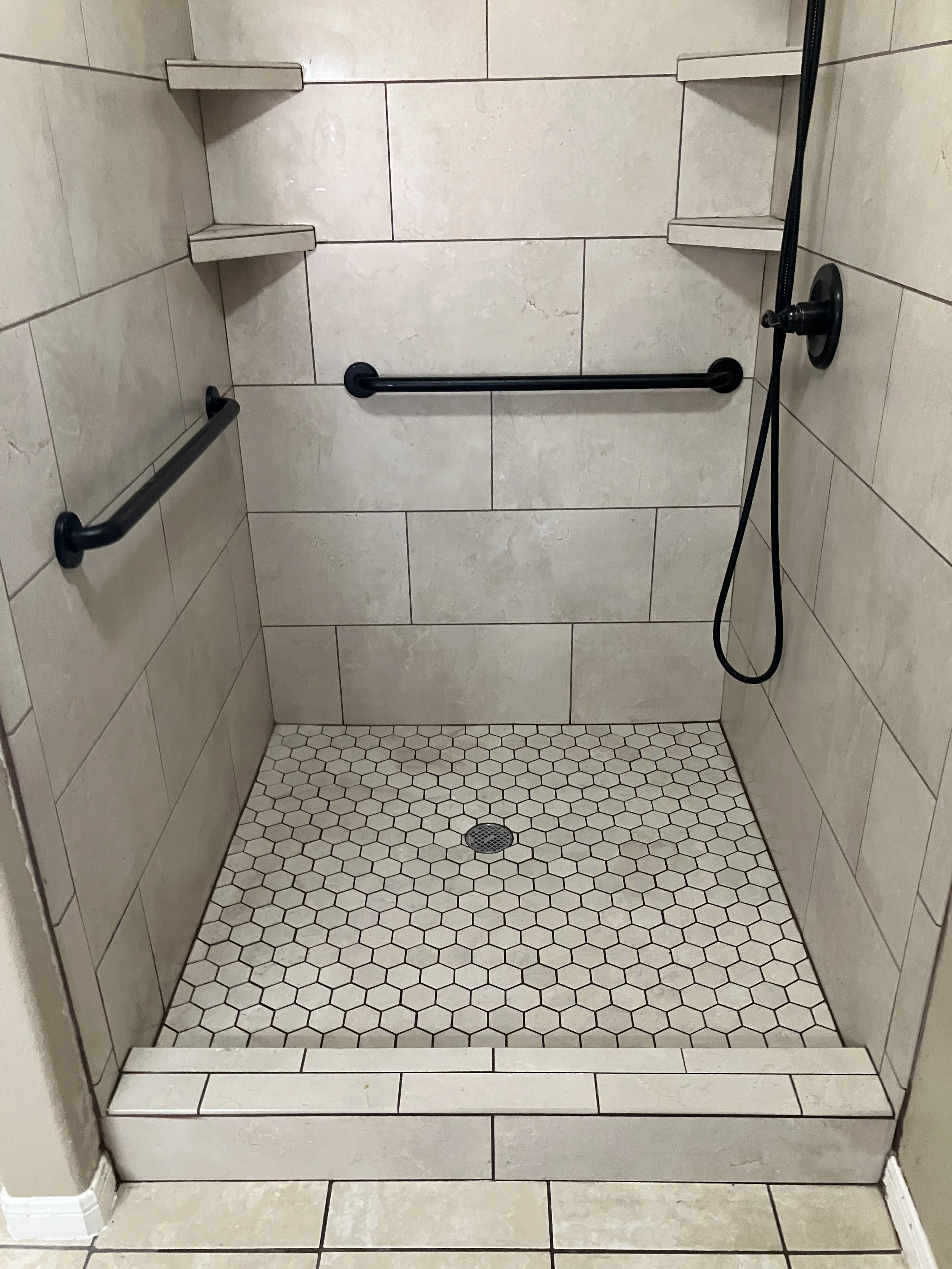 A walk-in shower with beige tiled walls, hexagonal tiled floor, two black grab bars, a black handheld showerhead on a hose, and two corner shelves.