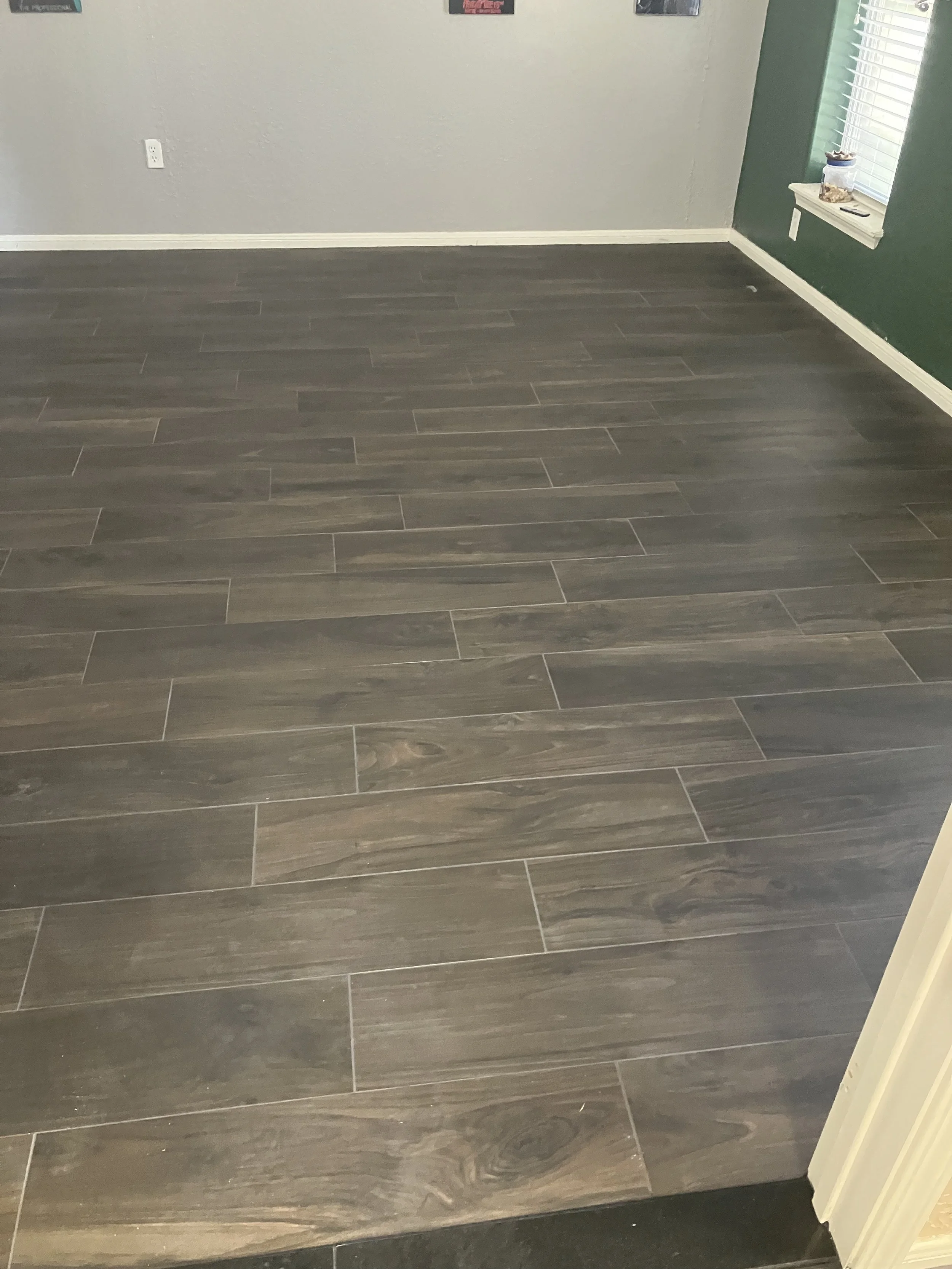 Interior of a room with wood-look tiled flooring, a gray wall, a green accent wall, a window with white blinds, and some framed pictures on the wall.