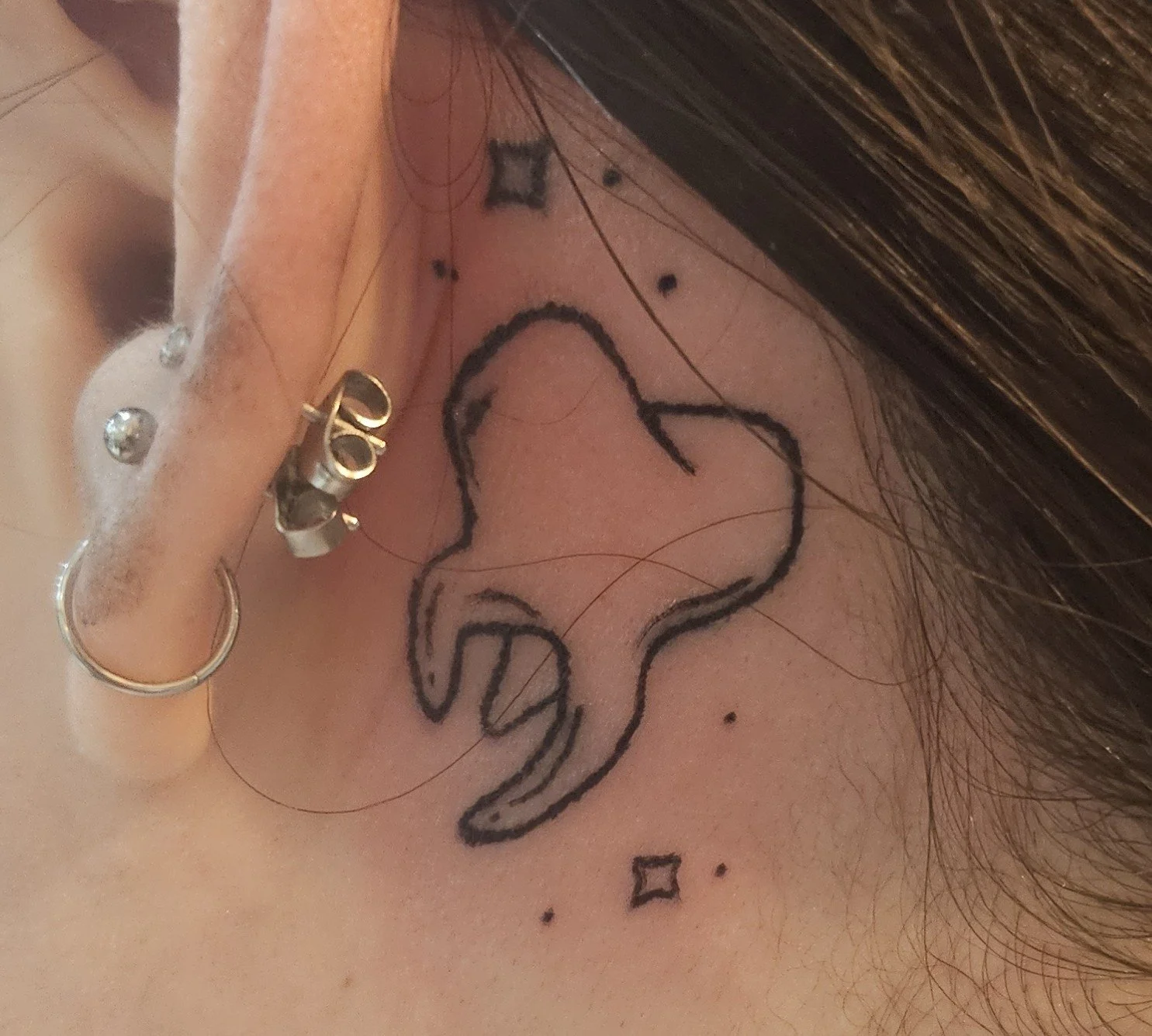 Close-up of a person's ear with multiple earrings and a tattoo of a tooth outline behind the ear, along with small star and diamond-shaped marks.