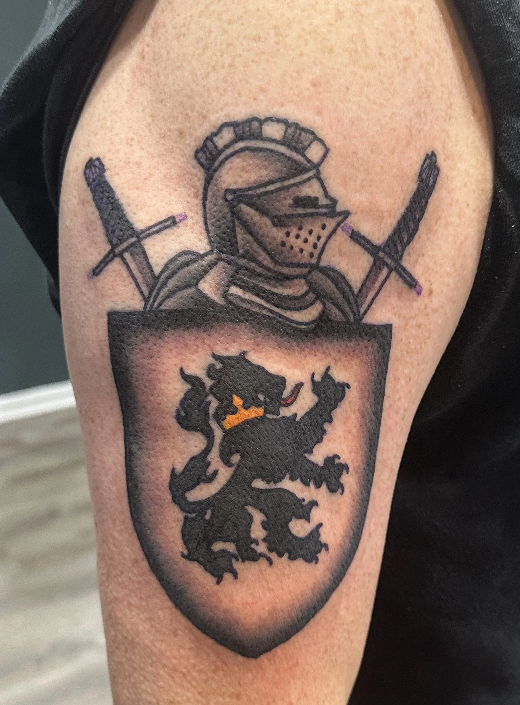 A tattoo featuring a medieval knight's helmet with crossed swords behind it, above a shield with a black lion with an orange tongue and collar.