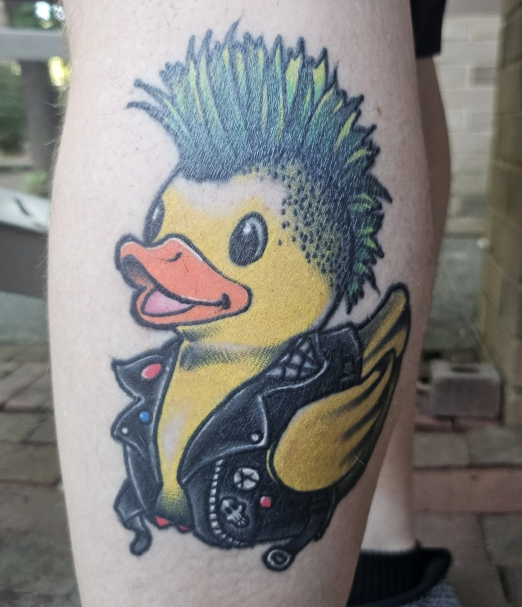 Tattoo of a yellow duck with a green mohawk hairstyle, wearing a black leather jacket, holding a video game controller.
