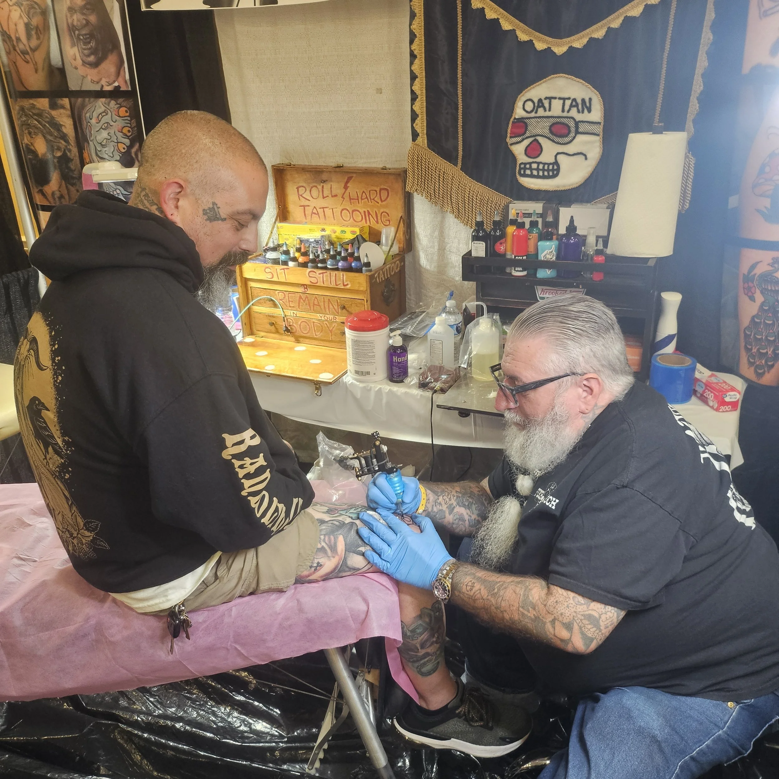 A tattoo artist is tattooing a man's leg while the man sits on a padded chair. The artist, wearing glasses and gloves, is focused on the tattoo, and the man appears to be relaxed. The background features various tattoo supplies, artwork, and signs re