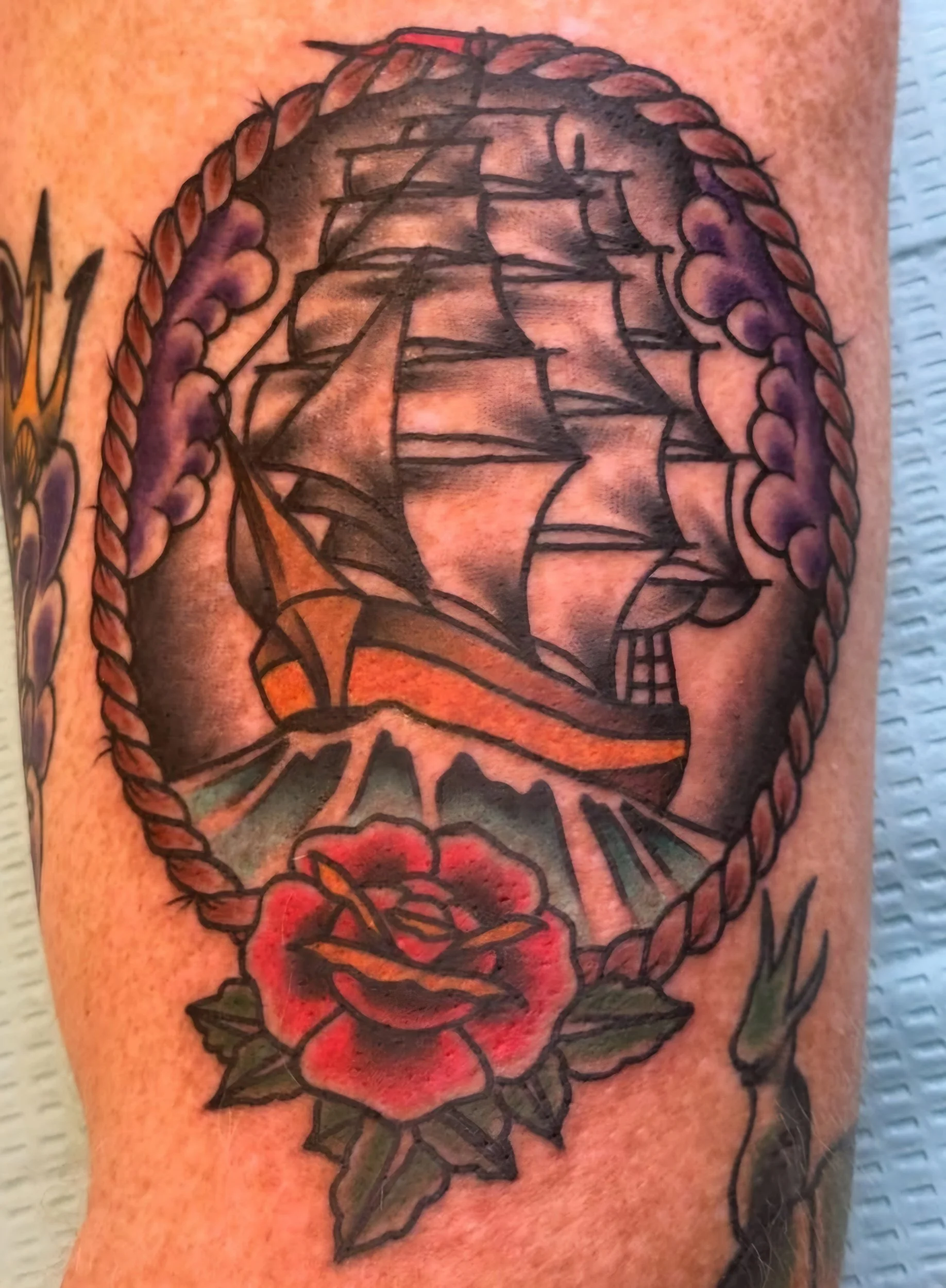 Tattoo of a sailing ship inside an oval rope frame, with a red rose and green leaves at the bottom.