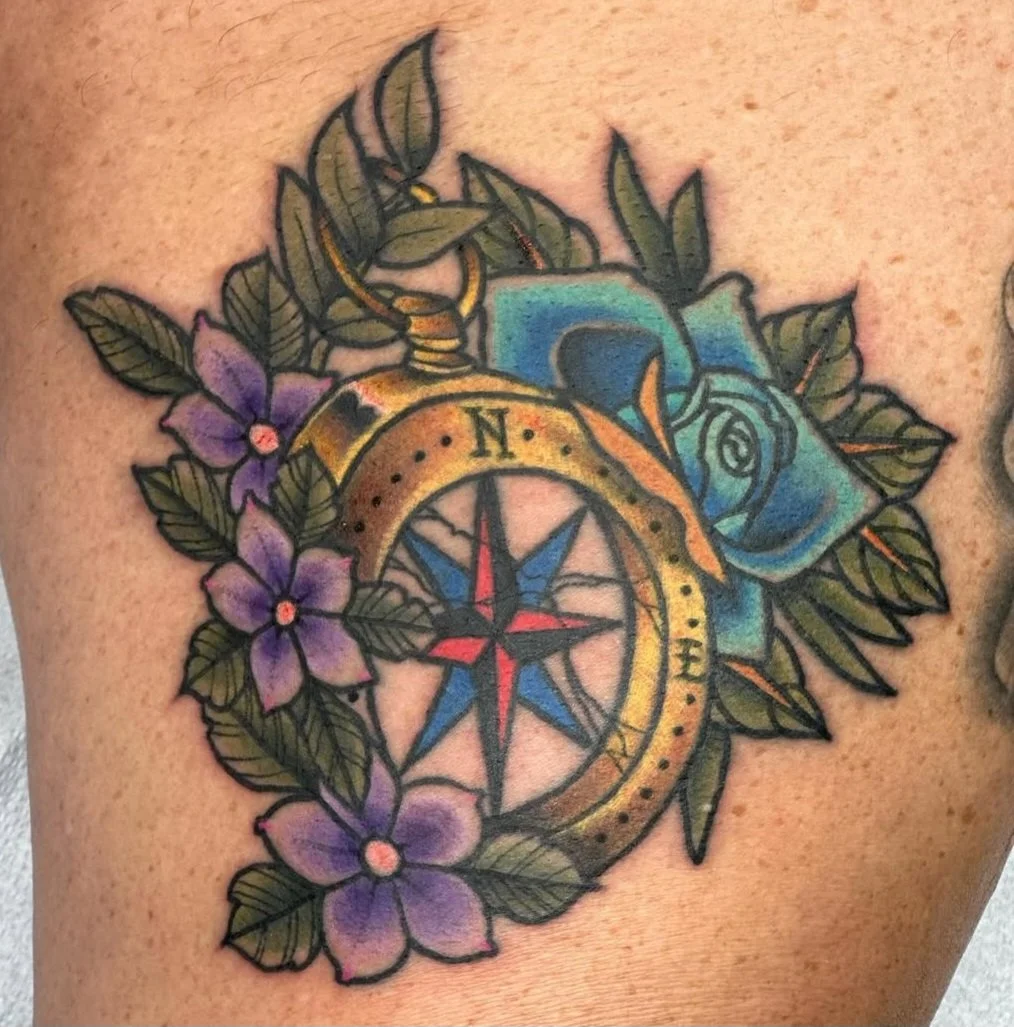 Tattoo of a compass, blue rose, purple flowers, and green leaves.