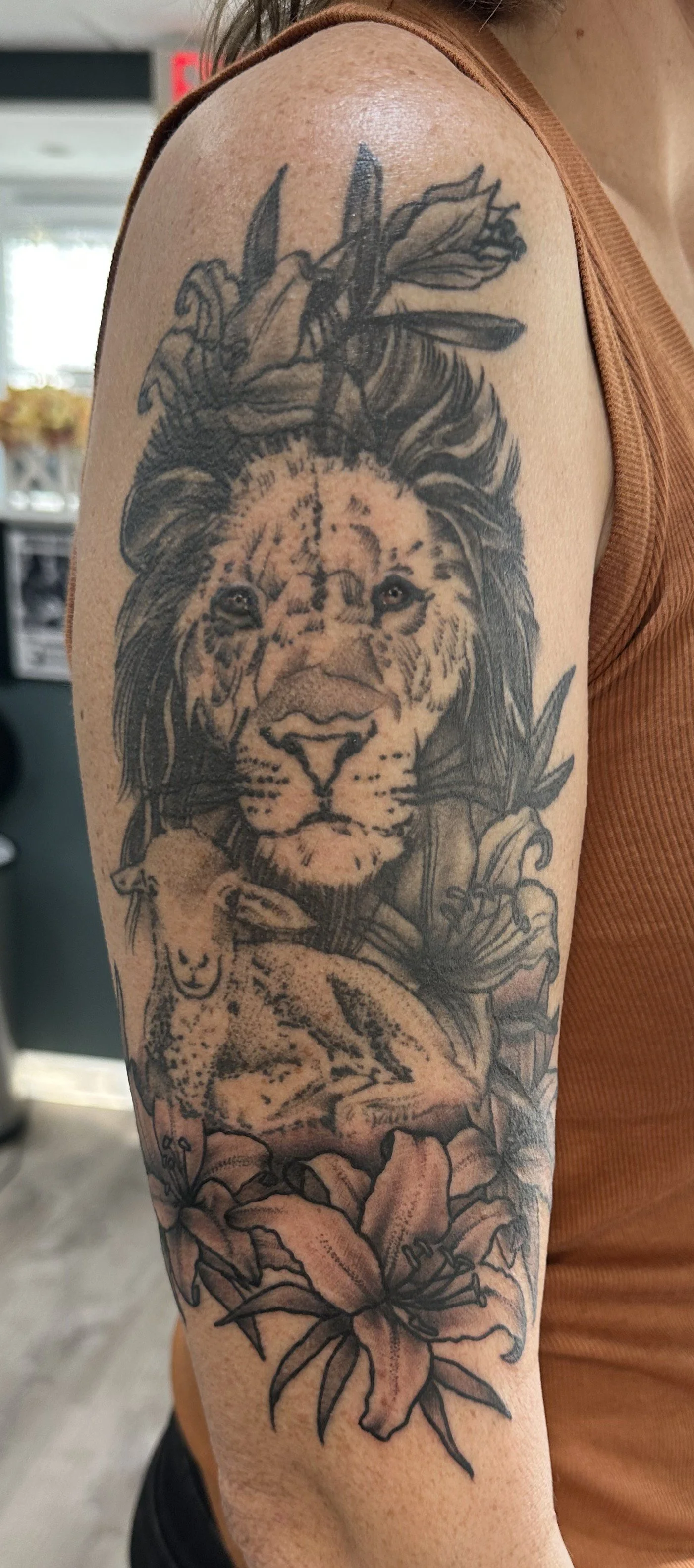 Tattoo of a lion's face with surrounding lilies, a bee flying above, and a small baby animal at the bottom, all in black and gray ink on an upper arm.